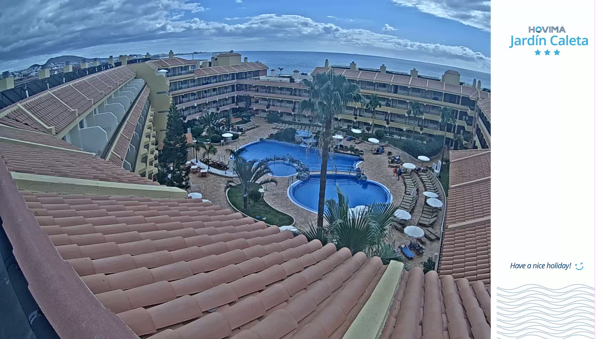 An aerial view of a resort complex with a large pool surrounded by buildings under a partly cloudy sky.