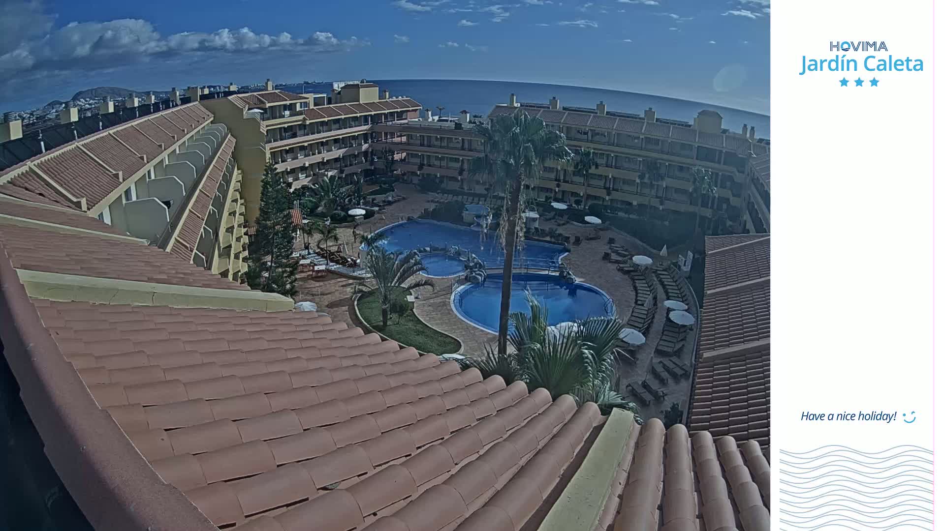 An aerial view shows a resort with a large, multi-level building surrounding a courtyard with two swimming pools, under a mostly sunny sky with some clouds.
