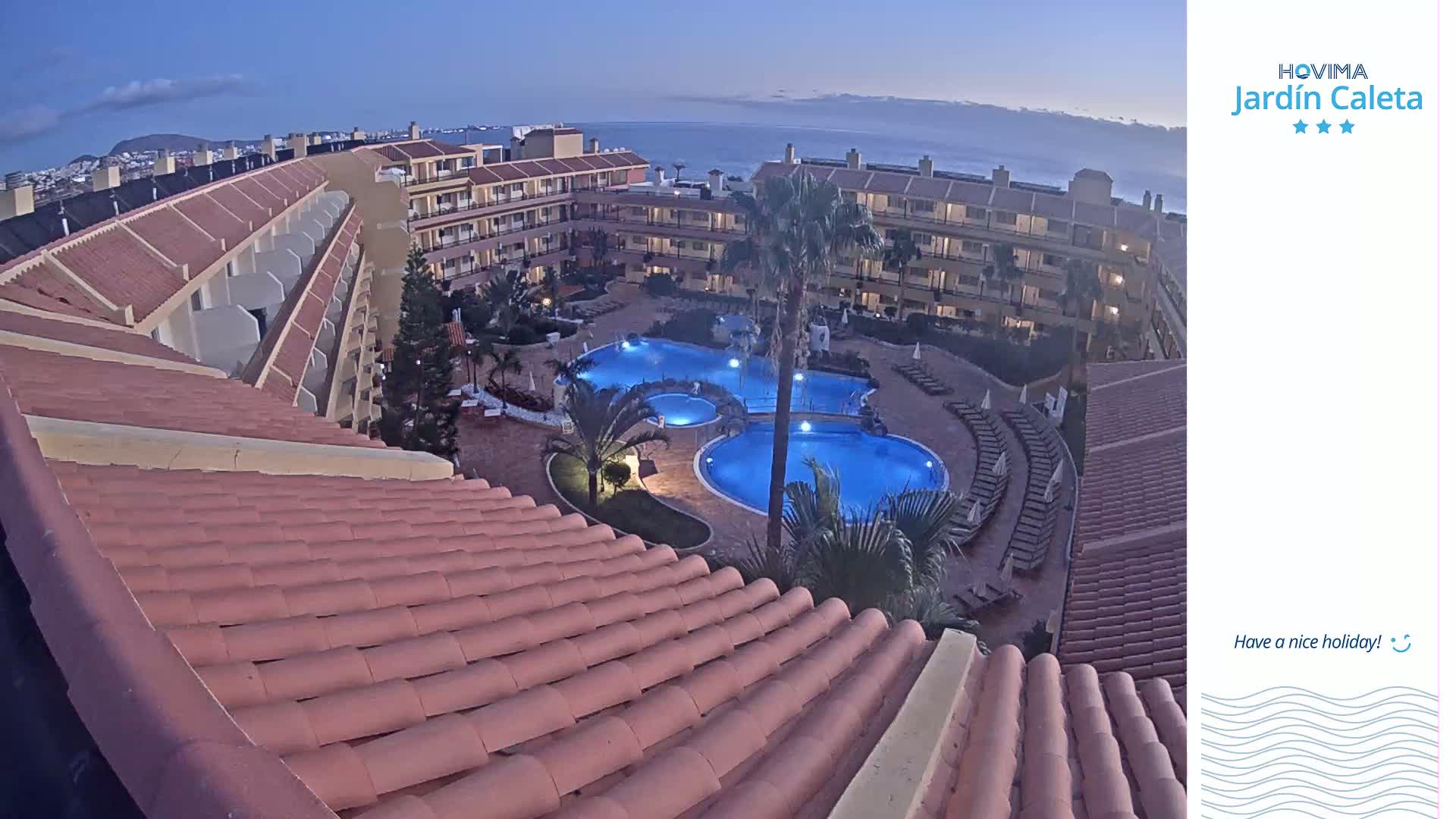 An illuminated resort pool complex is nestled within a low-rise building complex at dusk, overlooking a calm ocean.