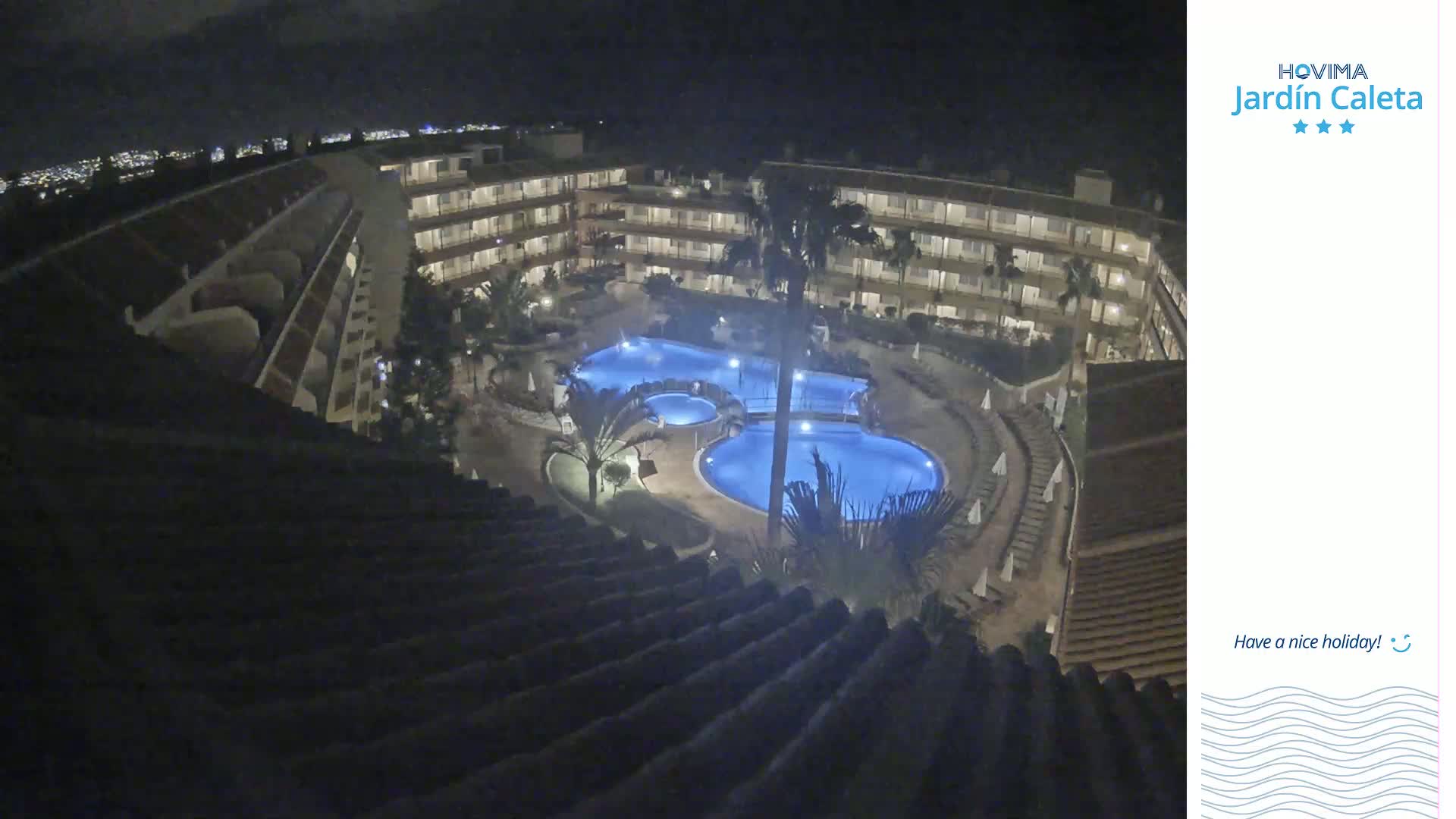 An illuminated resort pool area at night is viewed from an upper-level building.