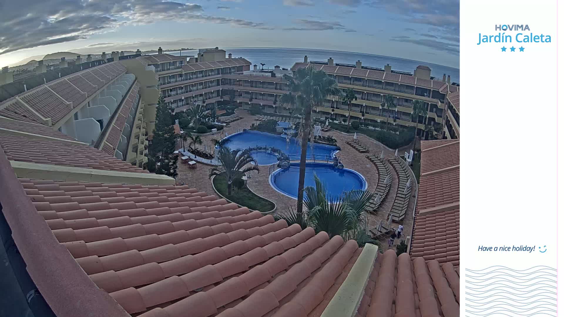 An overhead view shows a resort with a large, multi-sectioned pool surrounded by lounge chairs and buildings under a partly cloudy sky.