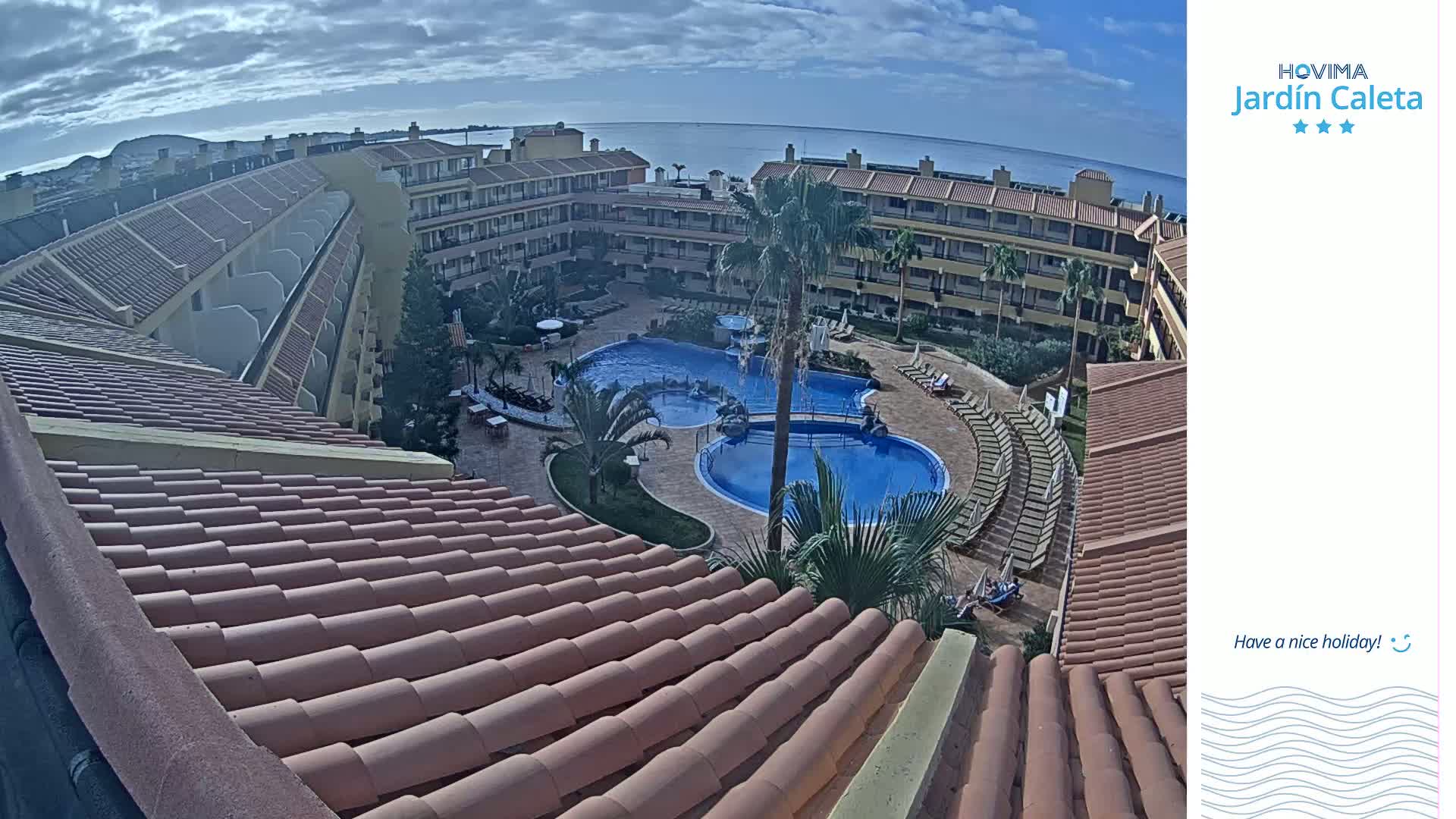 An overhead view shows a resort pool area with multiple buildings under a partly cloudy sky.