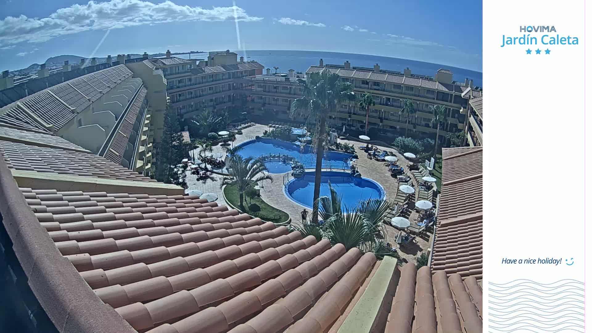 A rooftop view overlooking a sunny courtyard with two swimming pools, surrounded by a multi-story building and ocean in the distance.