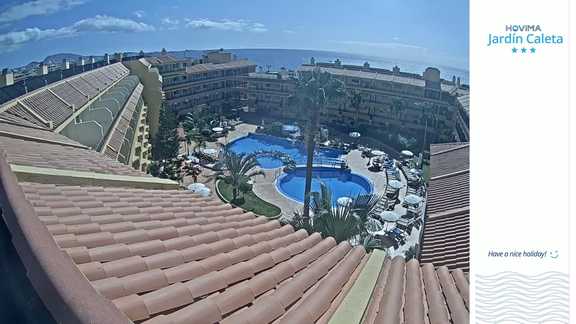 A sunny view from a rooftop shows a resort complex with multiple pools, surrounded by buildings and palm trees, overlooking a calm ocean.
