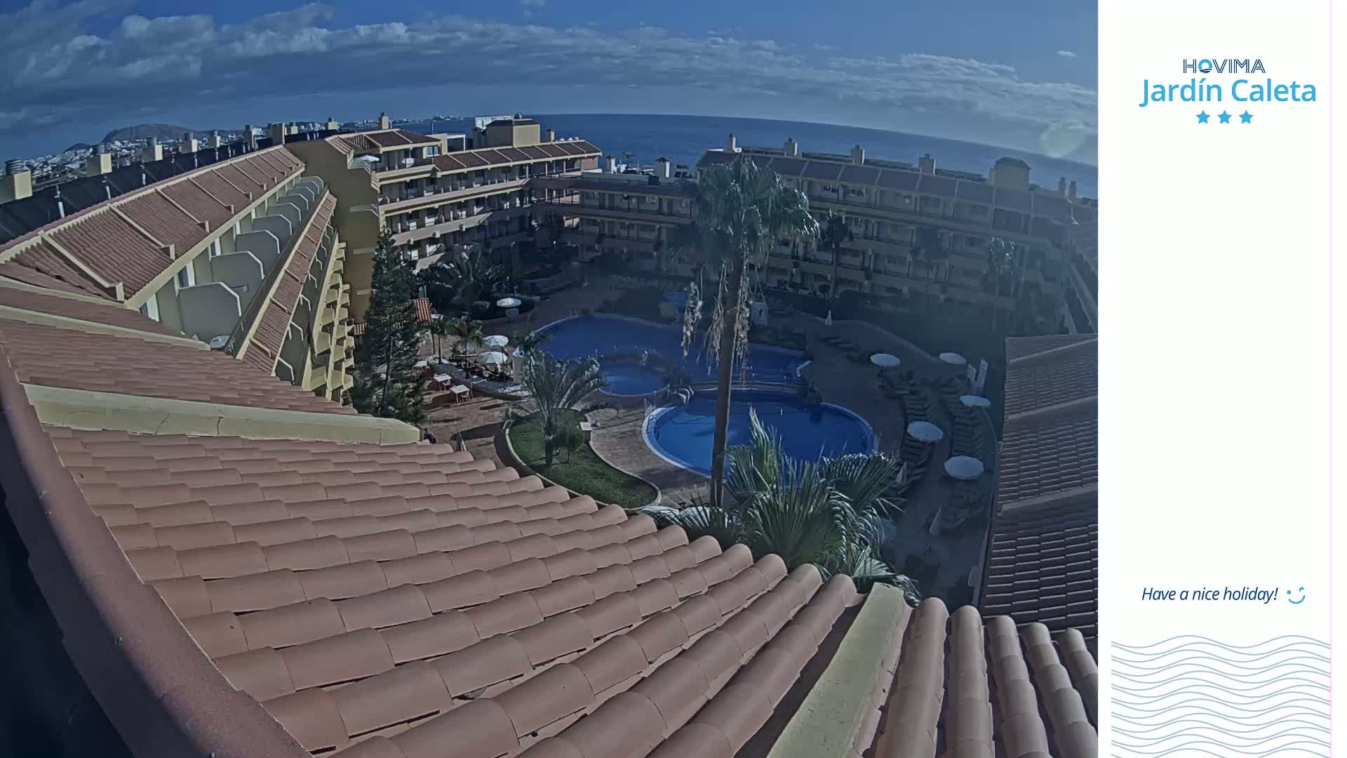 A partly cloudy day reveals a resort's courtyard with two swimming pools surrounded by buildings, viewed from an elevated perspective over a curved terracotta roof.