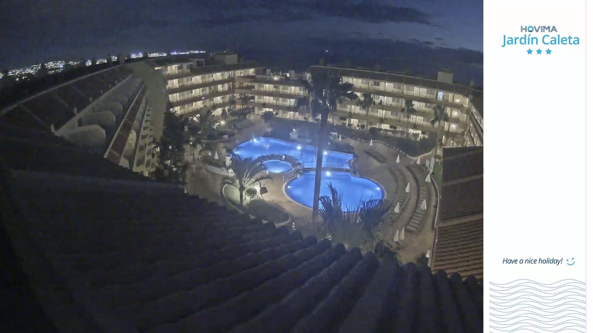 A nighttime aerial view shows a resort with several illuminated pools surrounded by buildings and palm trees under a dark sky.