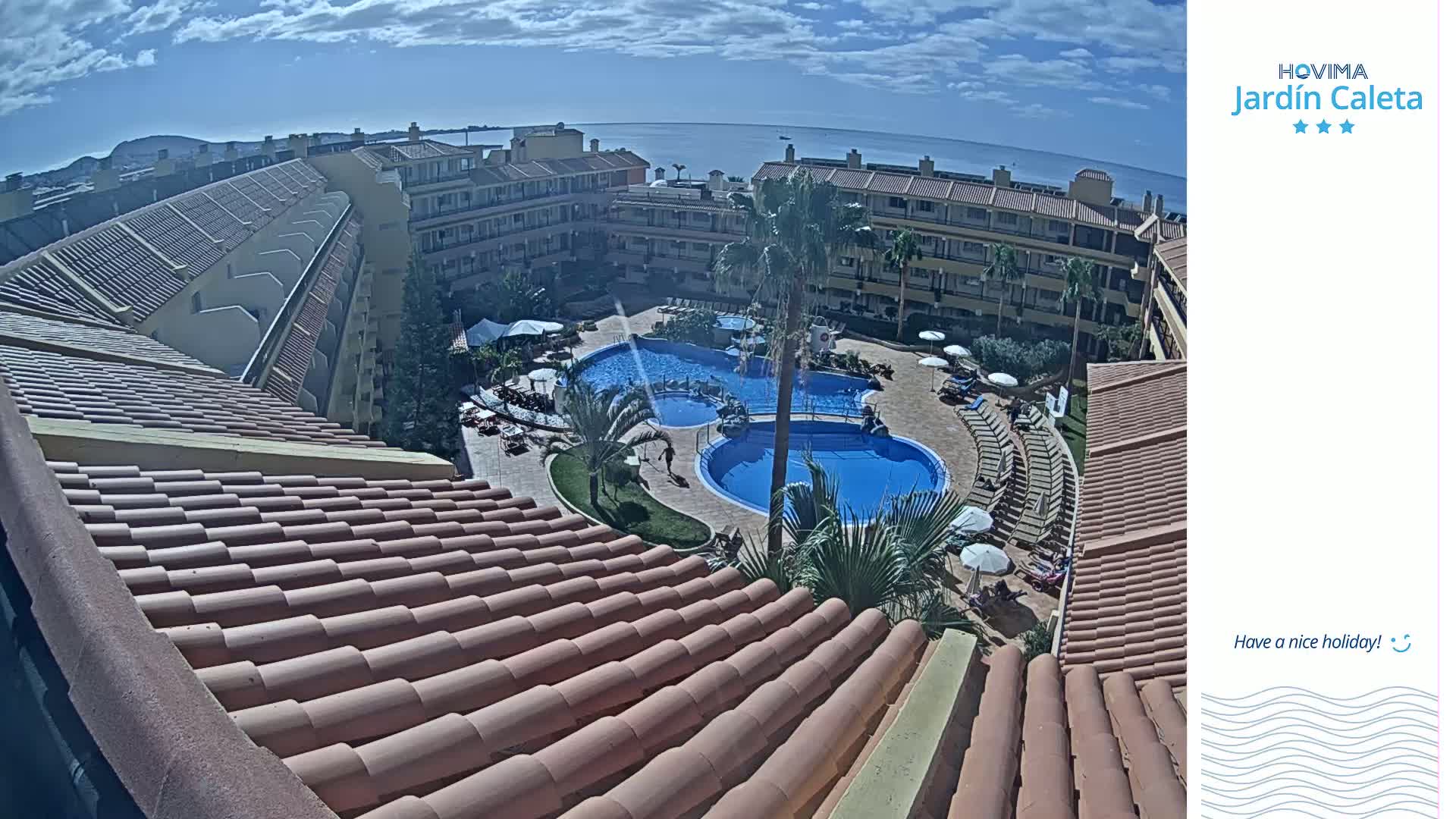 An overhead view of a resort complex with multiple pools surrounded by buildings, under a mostly sunny sky with a few clouds, overlooks the ocean in the distance.