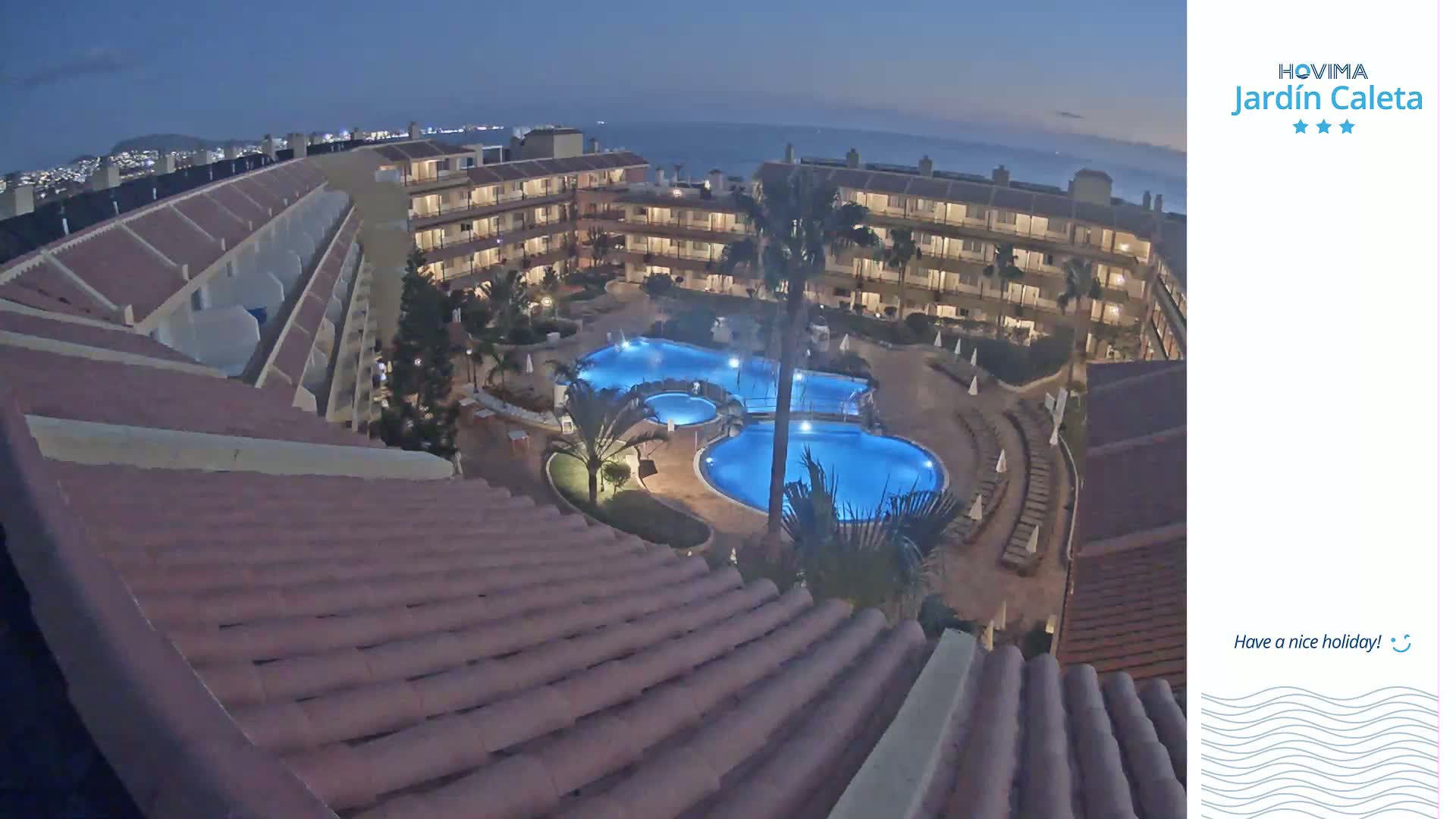An illuminated resort pool complex is nestled between multi-story buildings under a twilight sky.