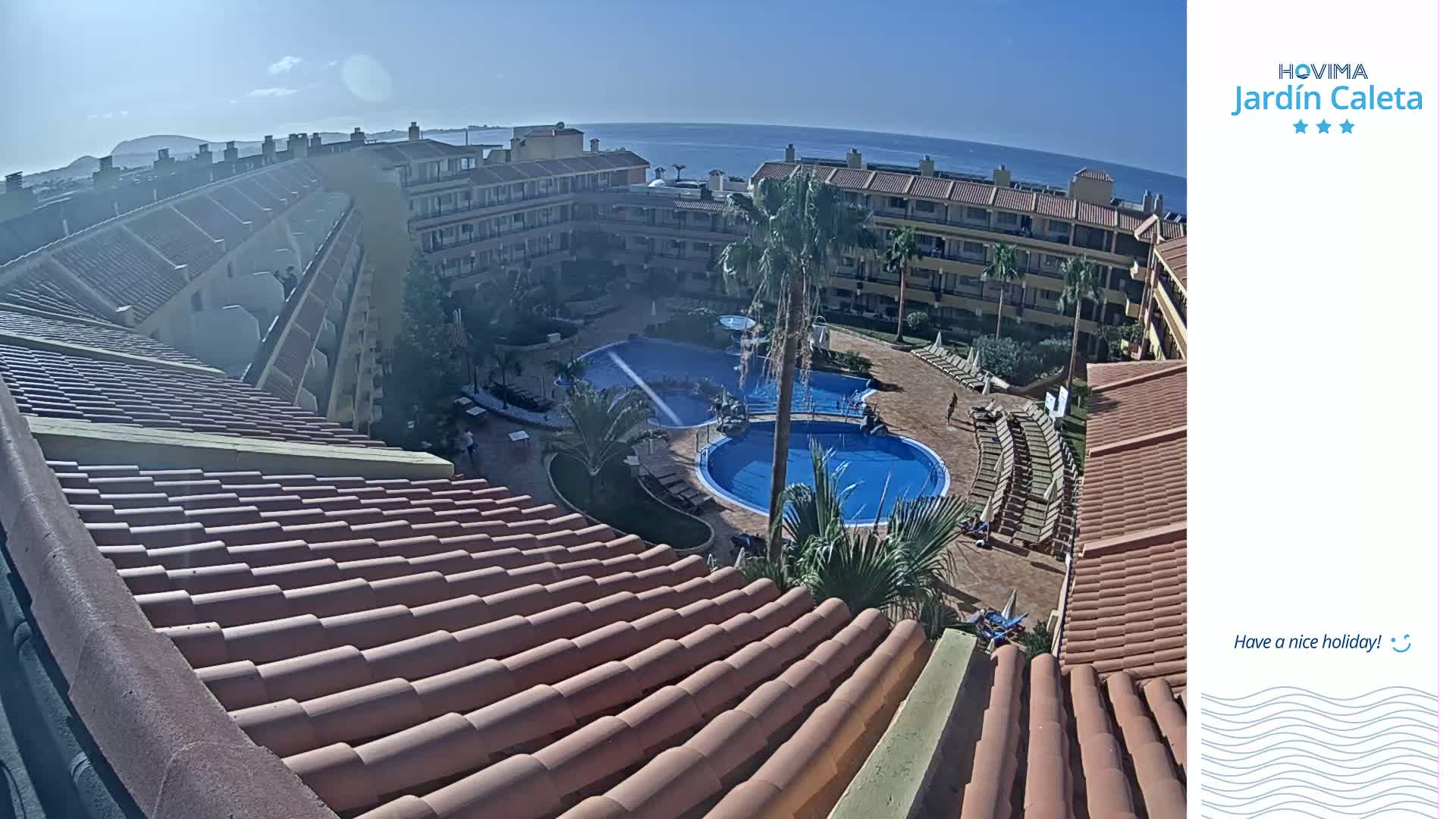 A sunny day overlooking a resort with multiple pools, surrounded by buildings with reddish-brown tile roofs and palm trees, all near a calm ocean.