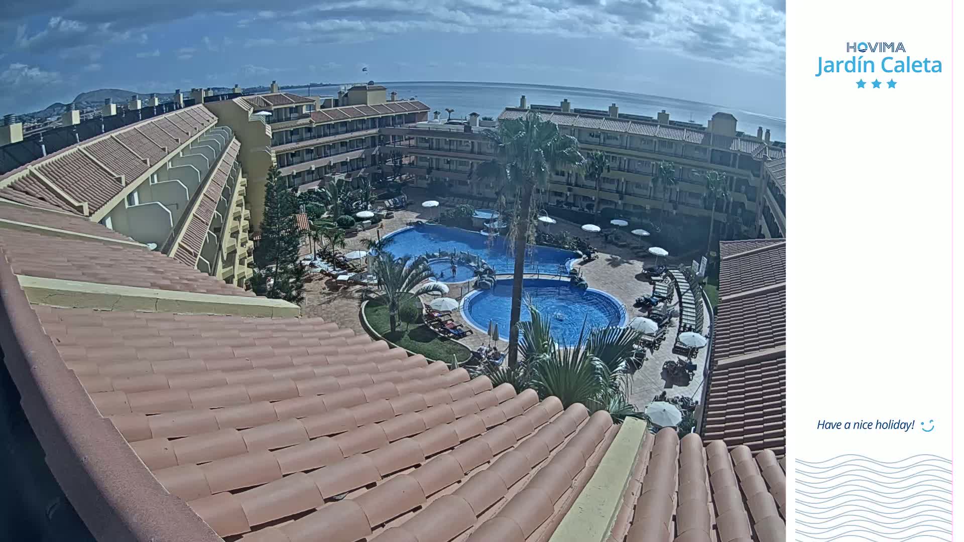 A tiled roof overlooks a sunny resort with two swimming pools surrounded by palm trees and lounge chairs, and a distant ocean view.