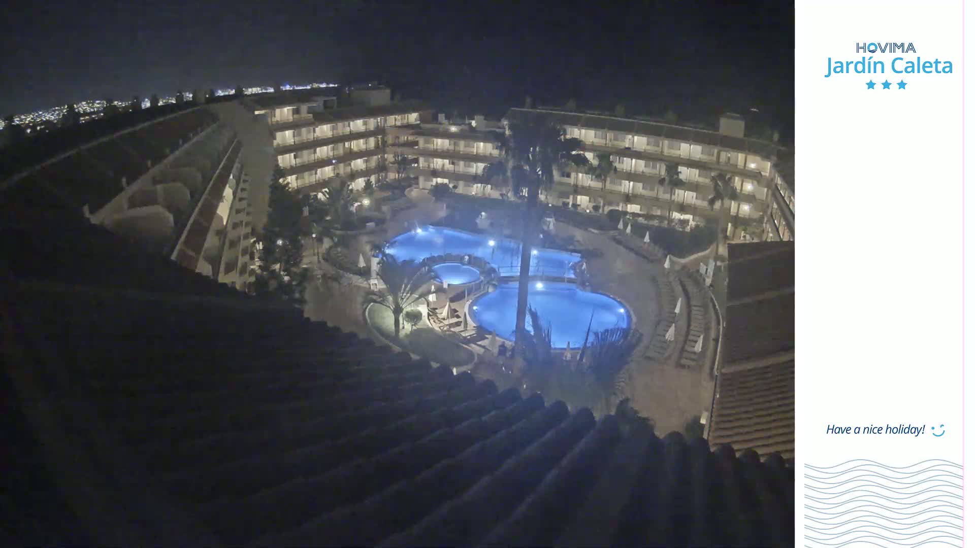 A nighttime aerial view shows a multi-story building complex surrounding illuminated swimming pools.