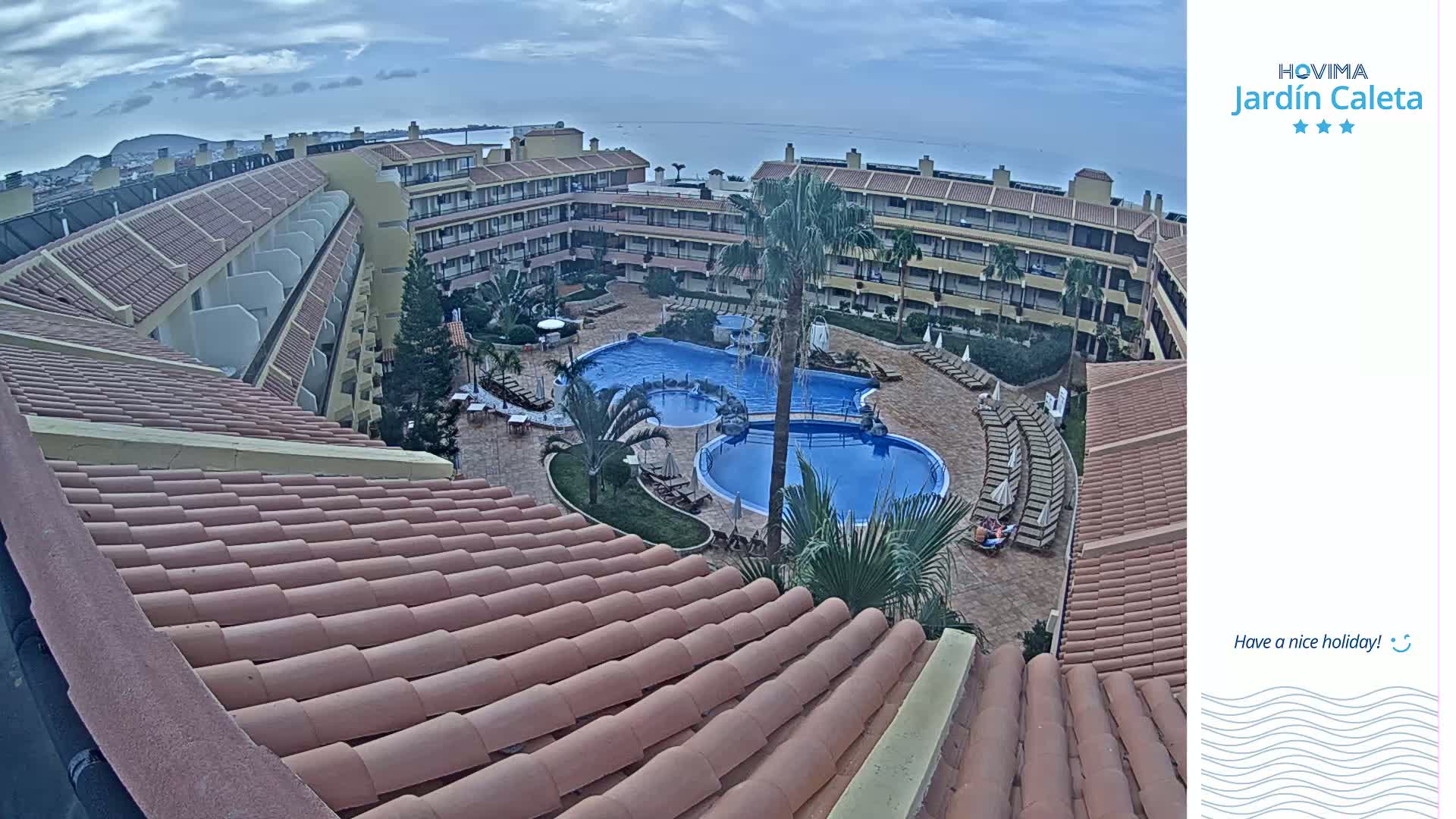 An elevated view of a resort courtyard with multiple pools, surrounded by buildings under a partly cloudy sky.