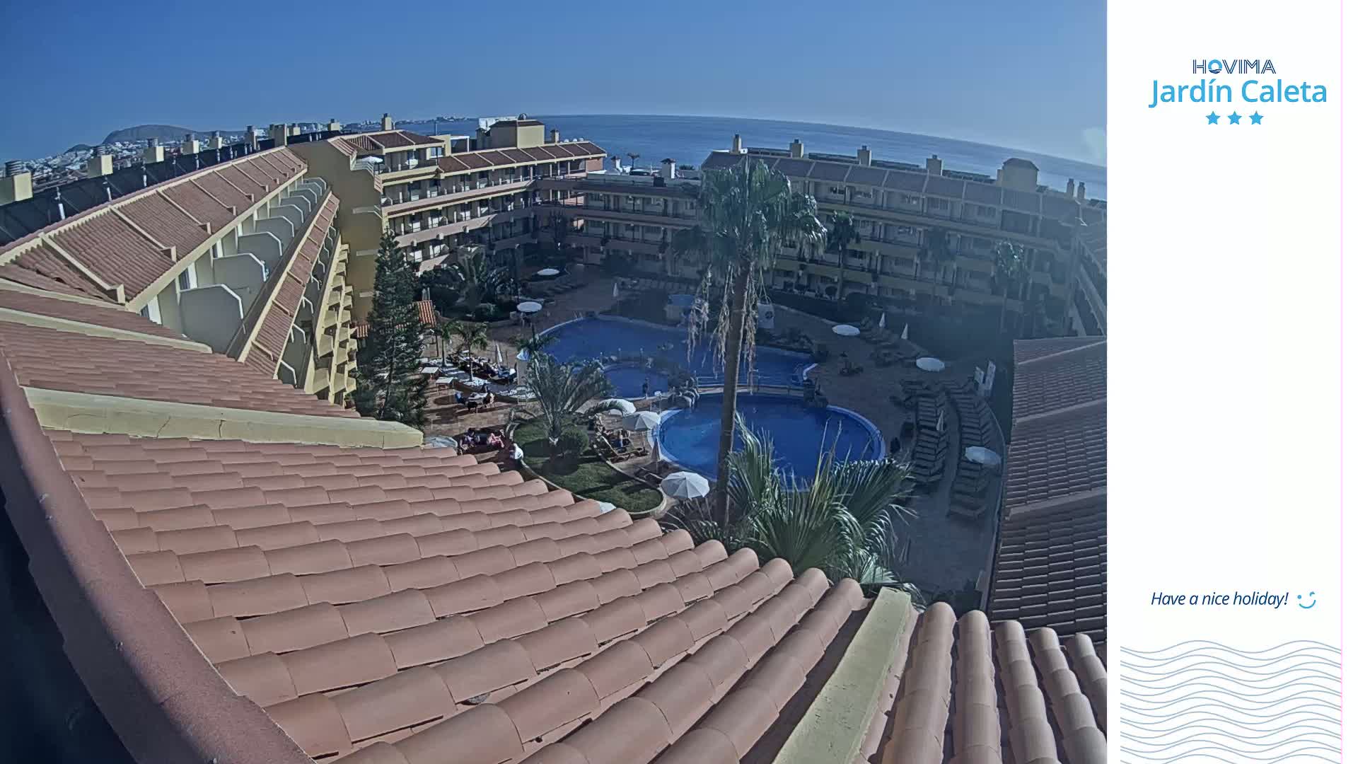 A tiled roof overlooks a resort pool area with multiple pools, palm trees, and buildings, all under a clear blue sky.