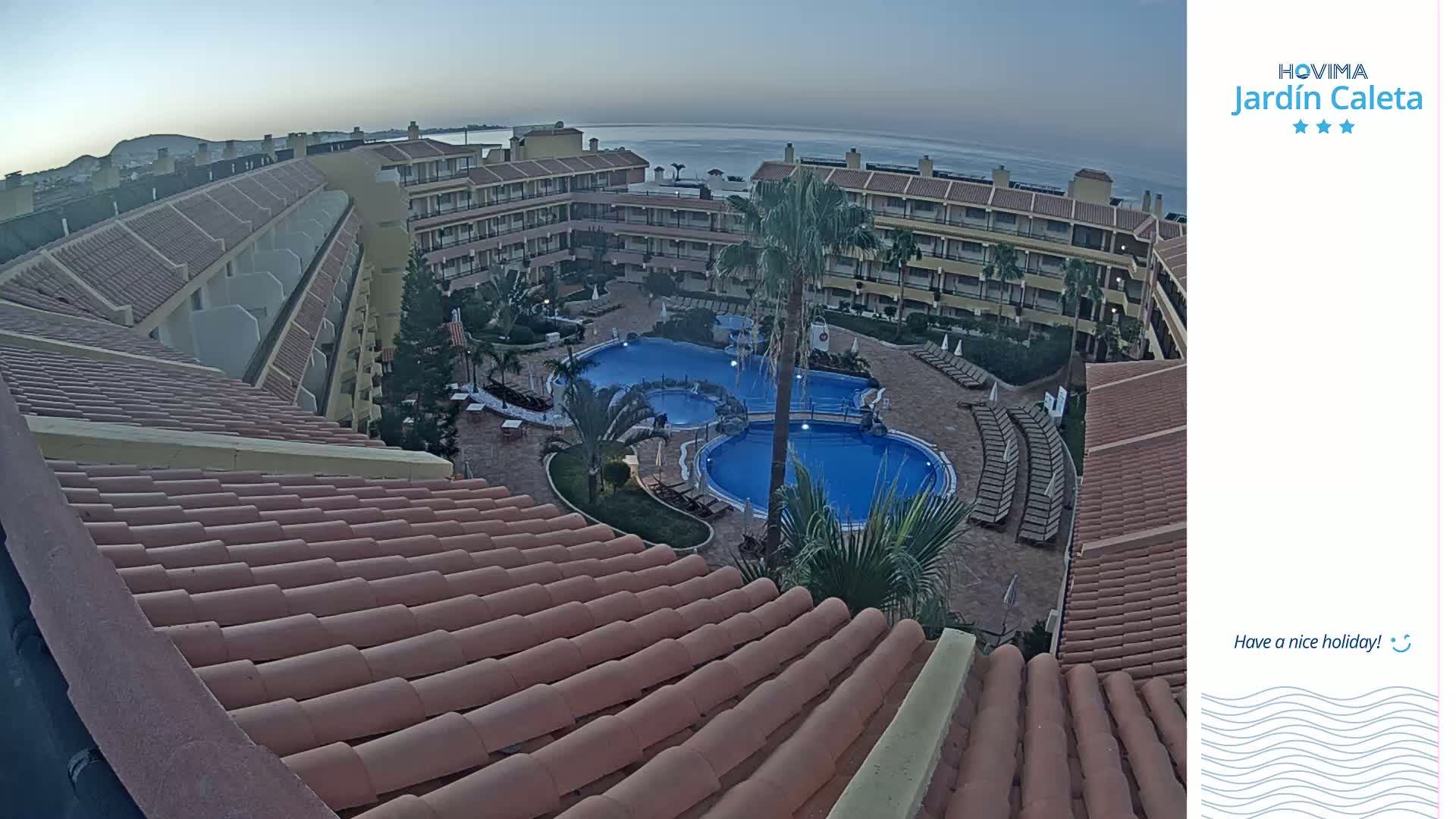 A tiled roof overlooks a courtyard with two swimming pools and a multi-story building complex at dusk under a clear sky.