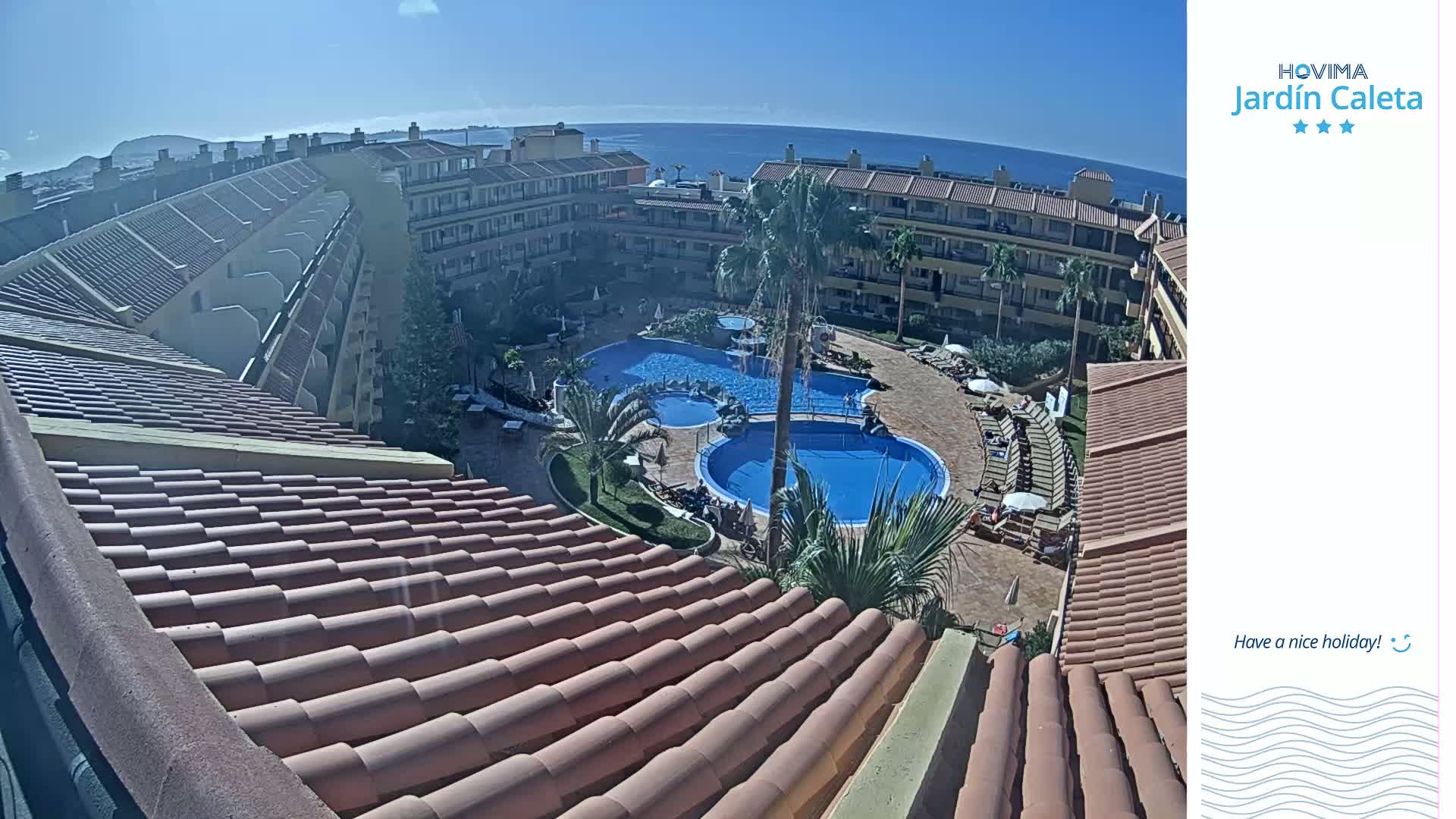 A sunny day overlooks a resort with multiple swimming pools surrounded by tan buildings and palm trees, all visible from a terracotta roof.