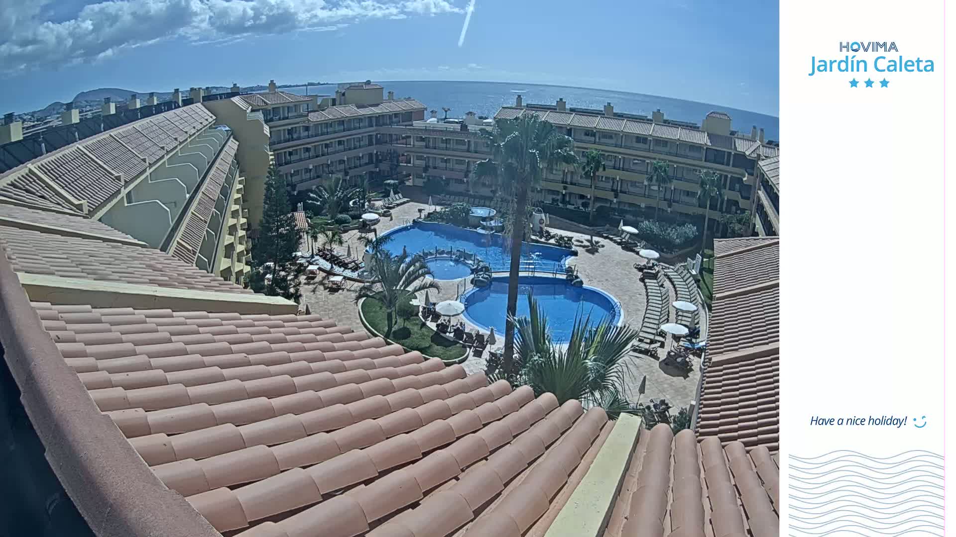 An overhead view of a resort complex with multiple pools, surrounded by buildings and palm trees, under a mostly sunny sky.