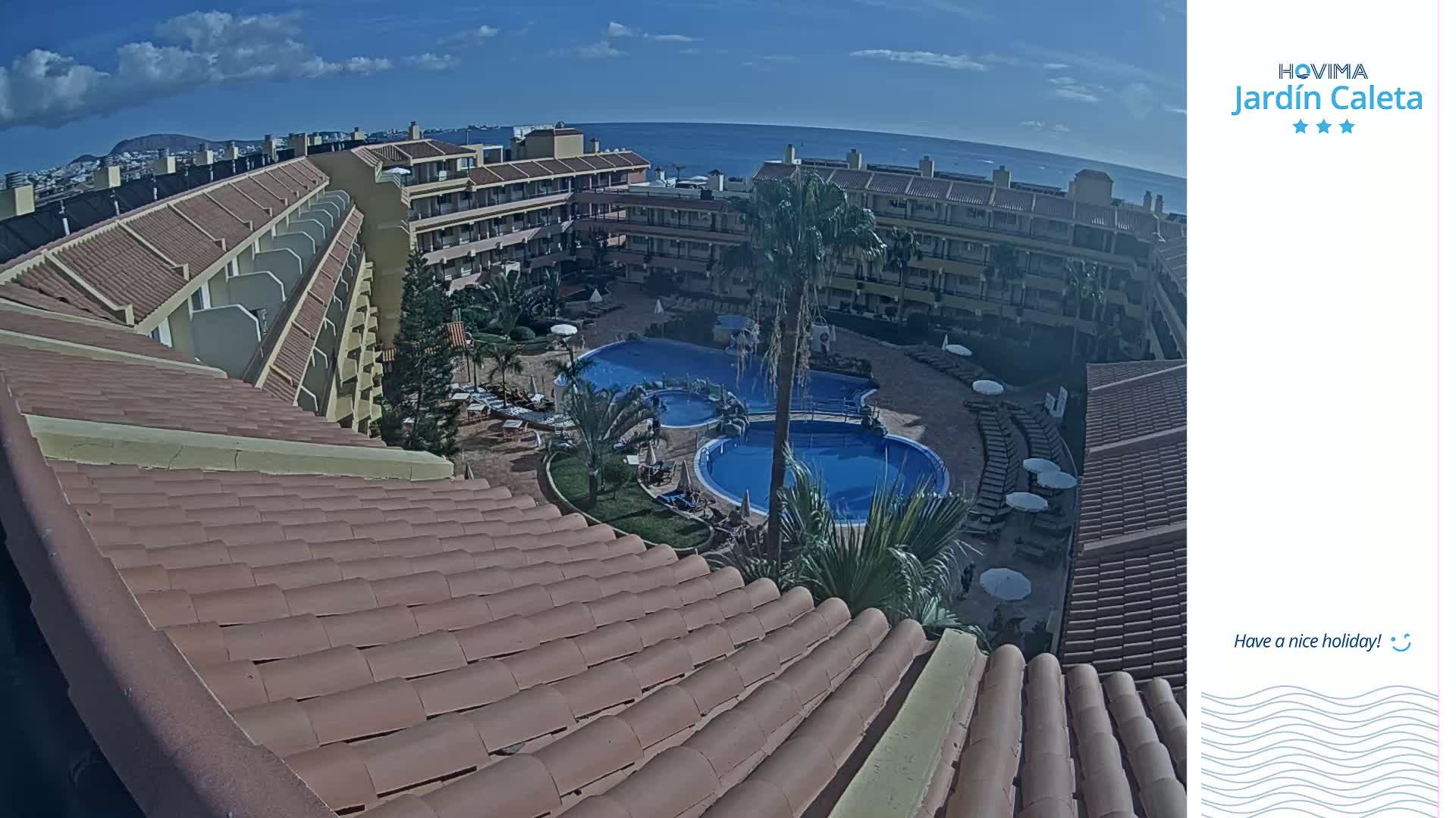 A view from a rooftop shows a resort with multiple buildings surrounding a swimming pool complex on a sunny day, with the ocean visible in the distance.