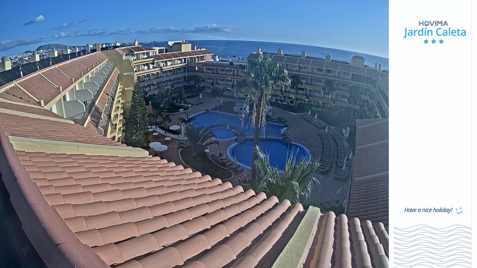 A curved terracotta roof overlooks a resort courtyard with multiple pools, palm trees, and a clear ocean view under a mostly sunny sky.