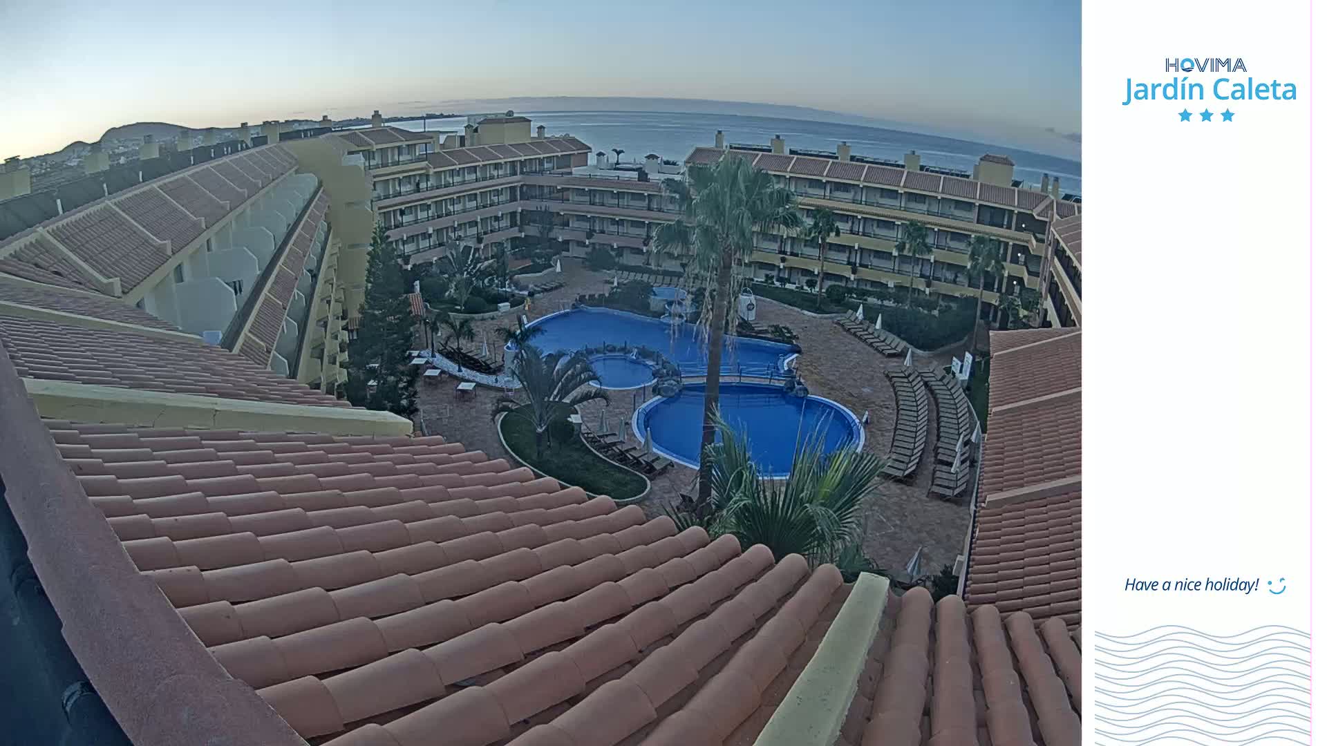 An aerial view of a resort complex with multiple buildings surrounding a swimming pool area, overlooking a calm ocean under a clear twilight sky.