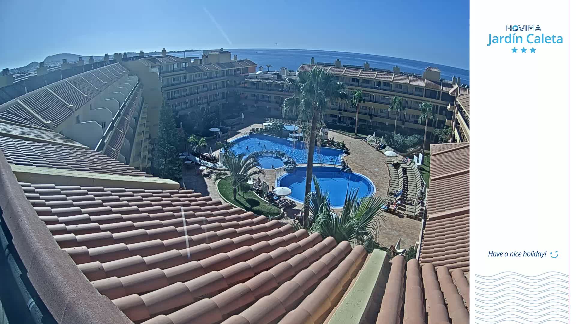 A high-angle view shows a resort pool area on a sunny day, with terracotta rooftops and the ocean visible in the background.