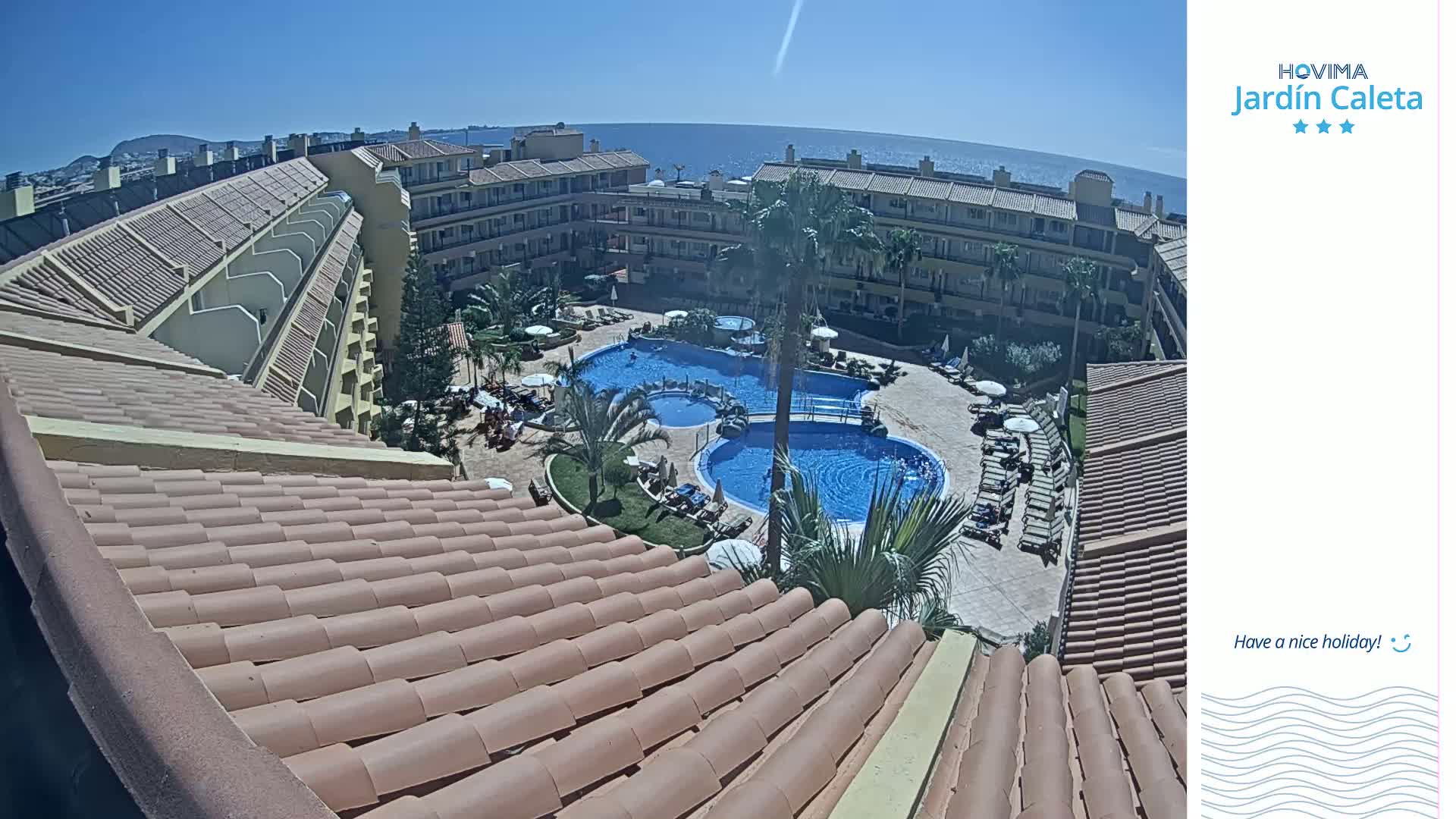 A tiled roof overlooks a sunny courtyard with two swimming pools, surrounded by a multi-story building and palm trees, next to a calm ocean.