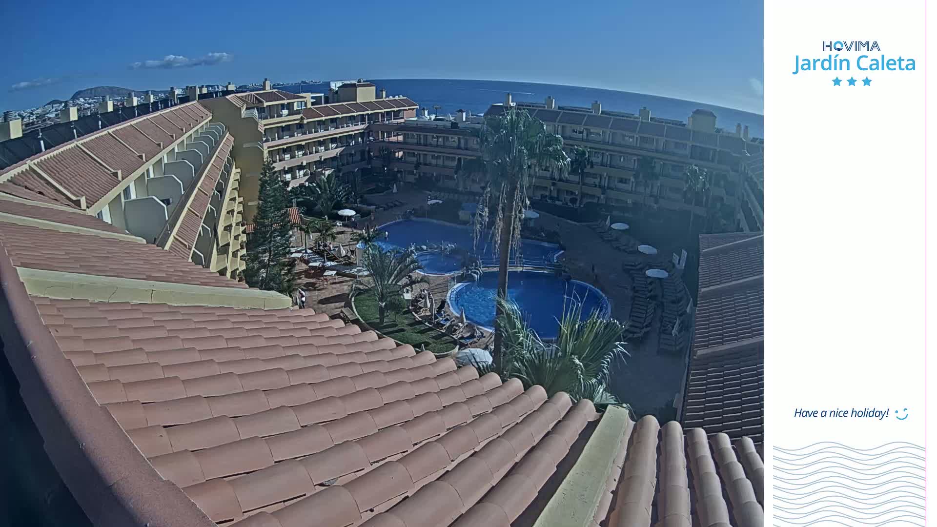 A sunny day overlooks a resort with two swimming pools surrounded by buildings and palm trees, and an ocean view in the distance.