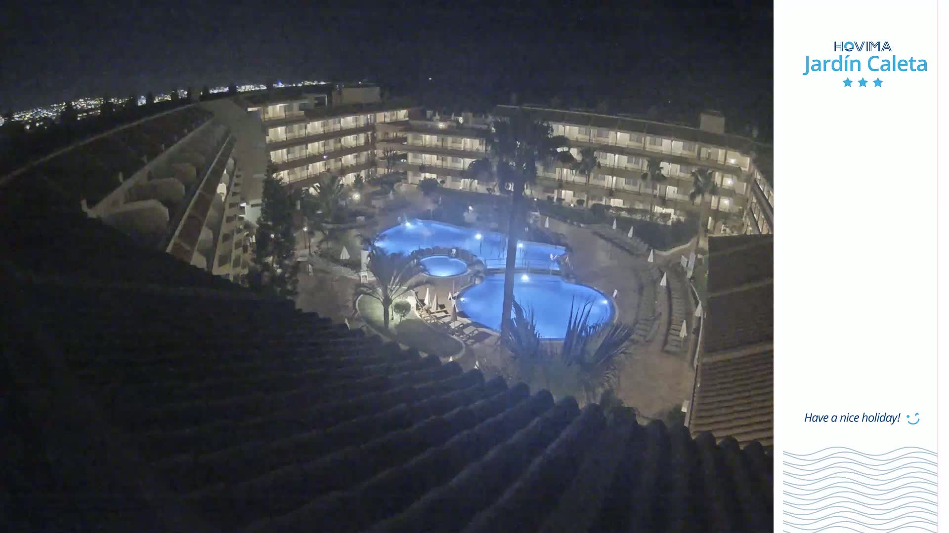 A nighttime view from a rooftop shows a multi-story building complex surrounding a pool lit with blue lights.
