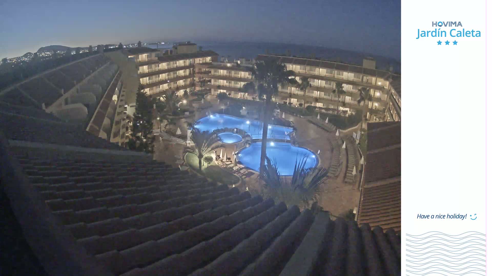 An illuminated resort pool complex is situated at night within a multi-story building, overlooking a dark coastline under a clear night sky.