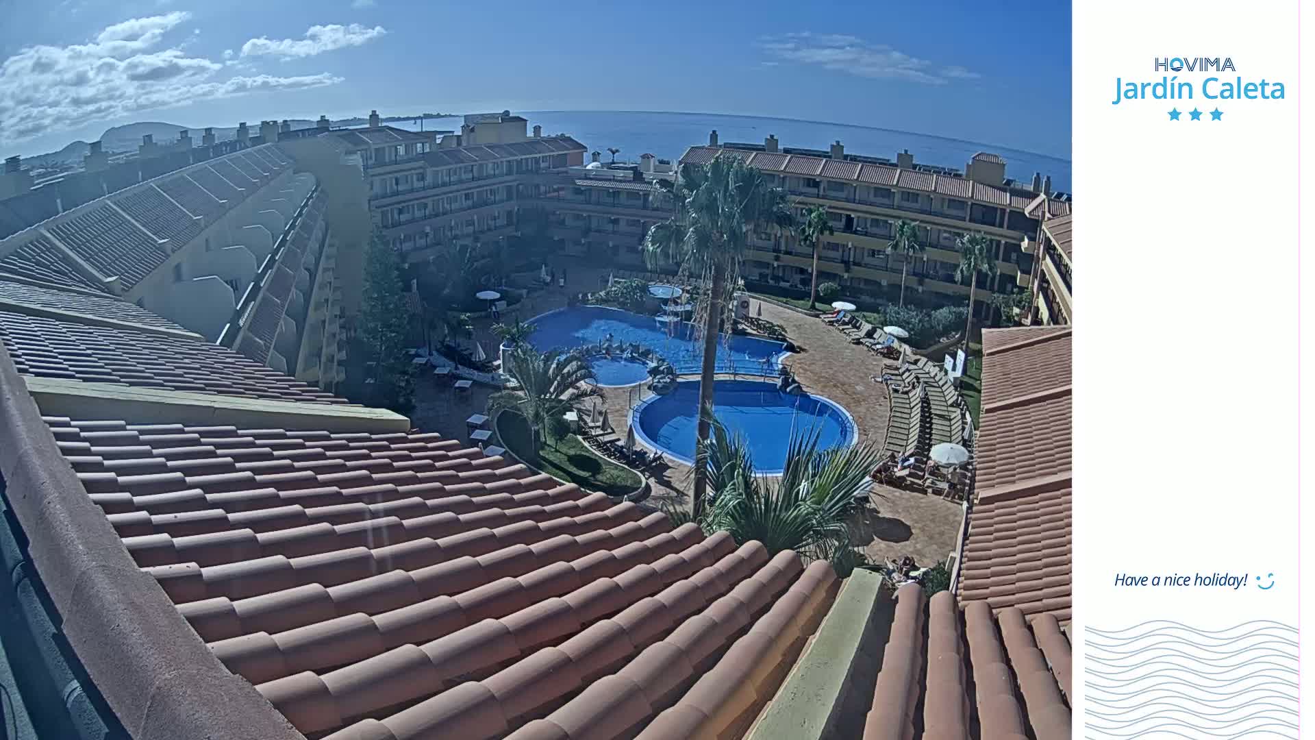 An aerial view shows a resort courtyard with multiple swimming pools, surrounded by buildings under a sunny sky with a calm ocean visible in the distance.