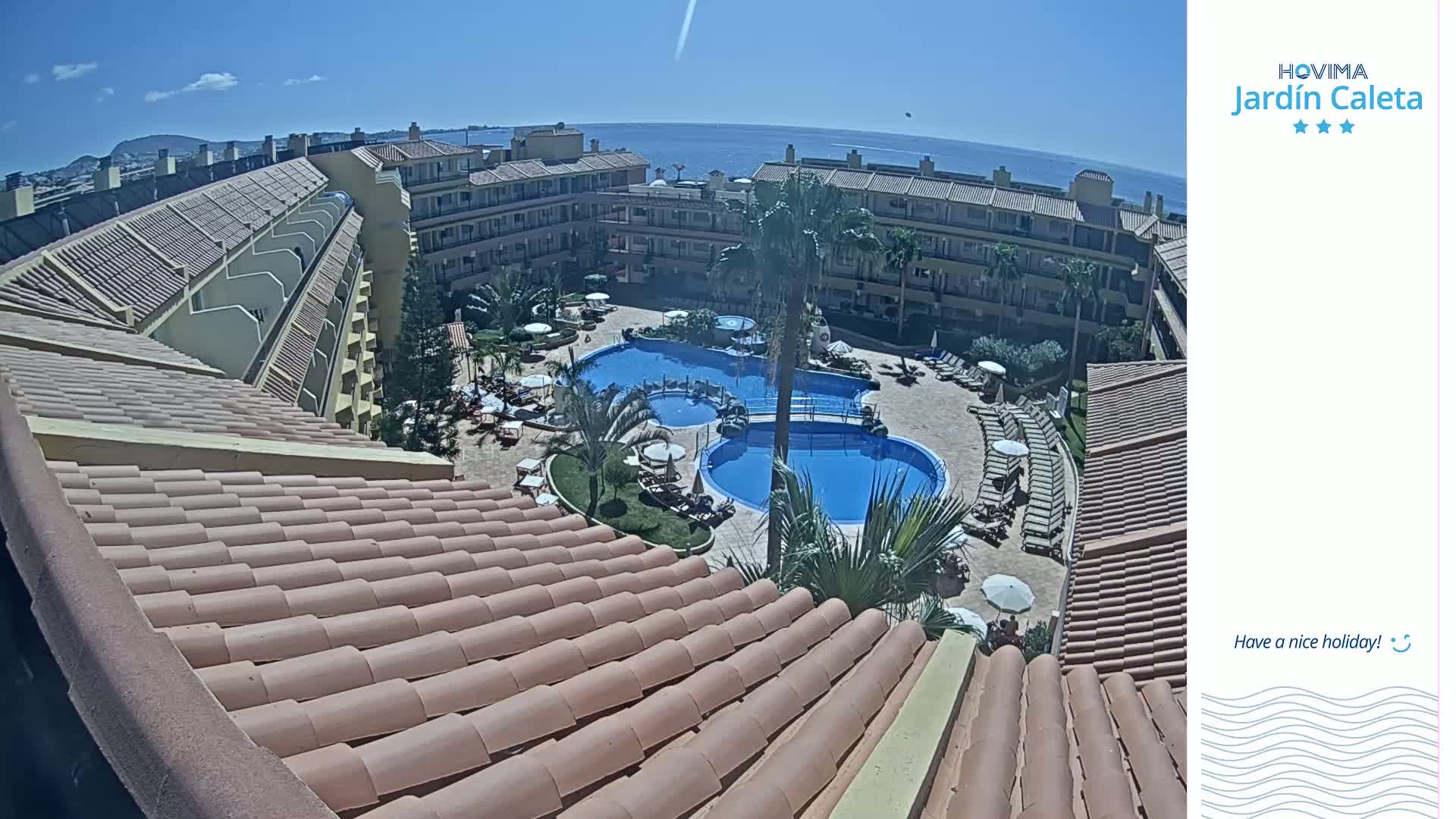 A rooftop view overlooking a sunny resort with multiple swimming pools surrounded by lounge chairs and buildings, a clear blue ocean visible in the distance.