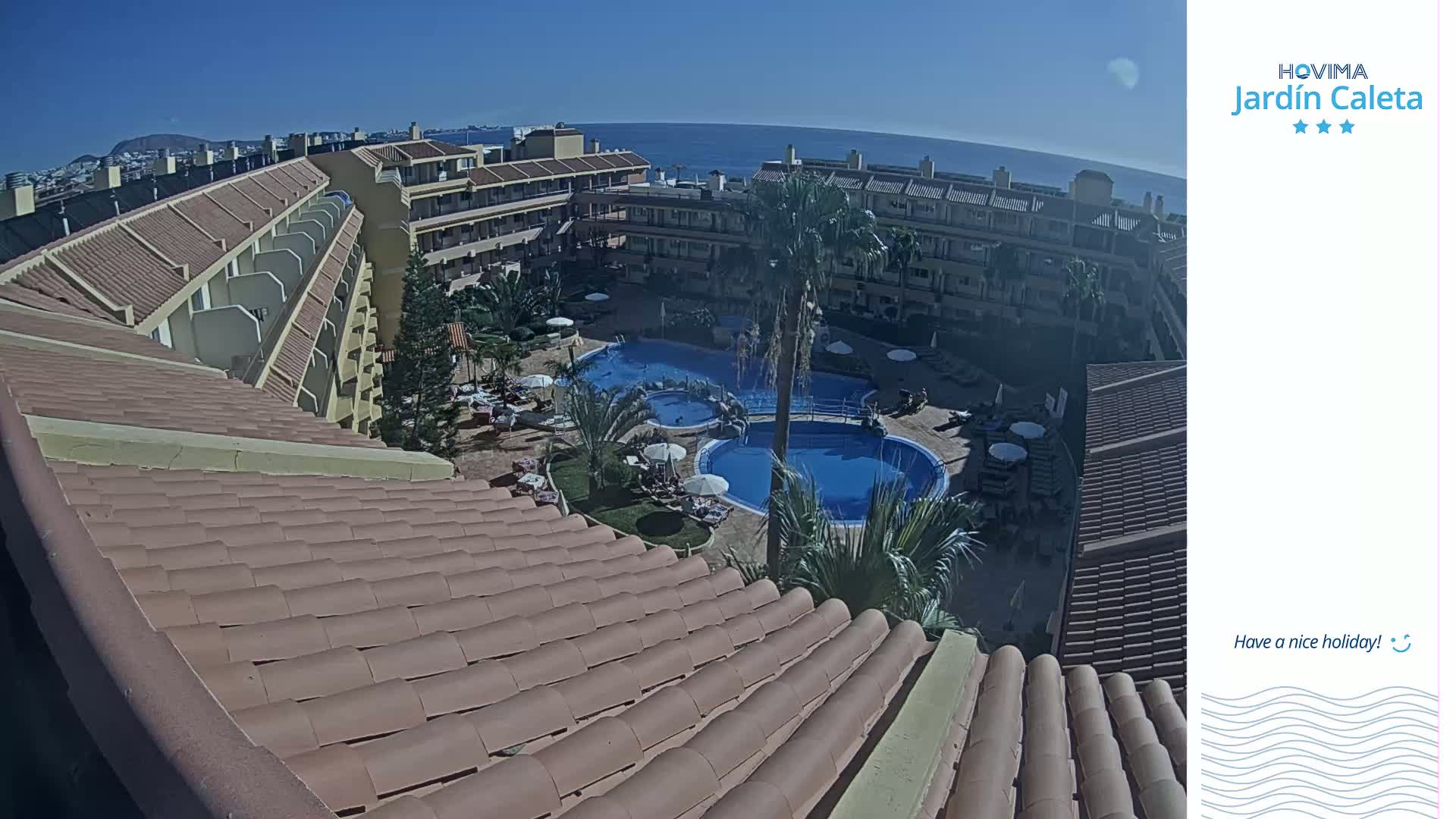 An aerial view of a resort with two swimming pools, surrounded by buildings under a clear sunny sky.