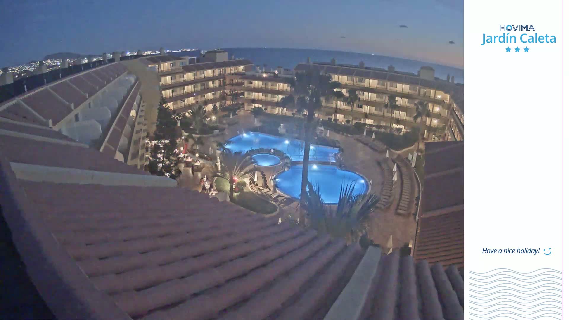 An aerial nighttime view shows a resort complex with multiple pools illuminated, surrounded by buildings under a clear, dark sky.