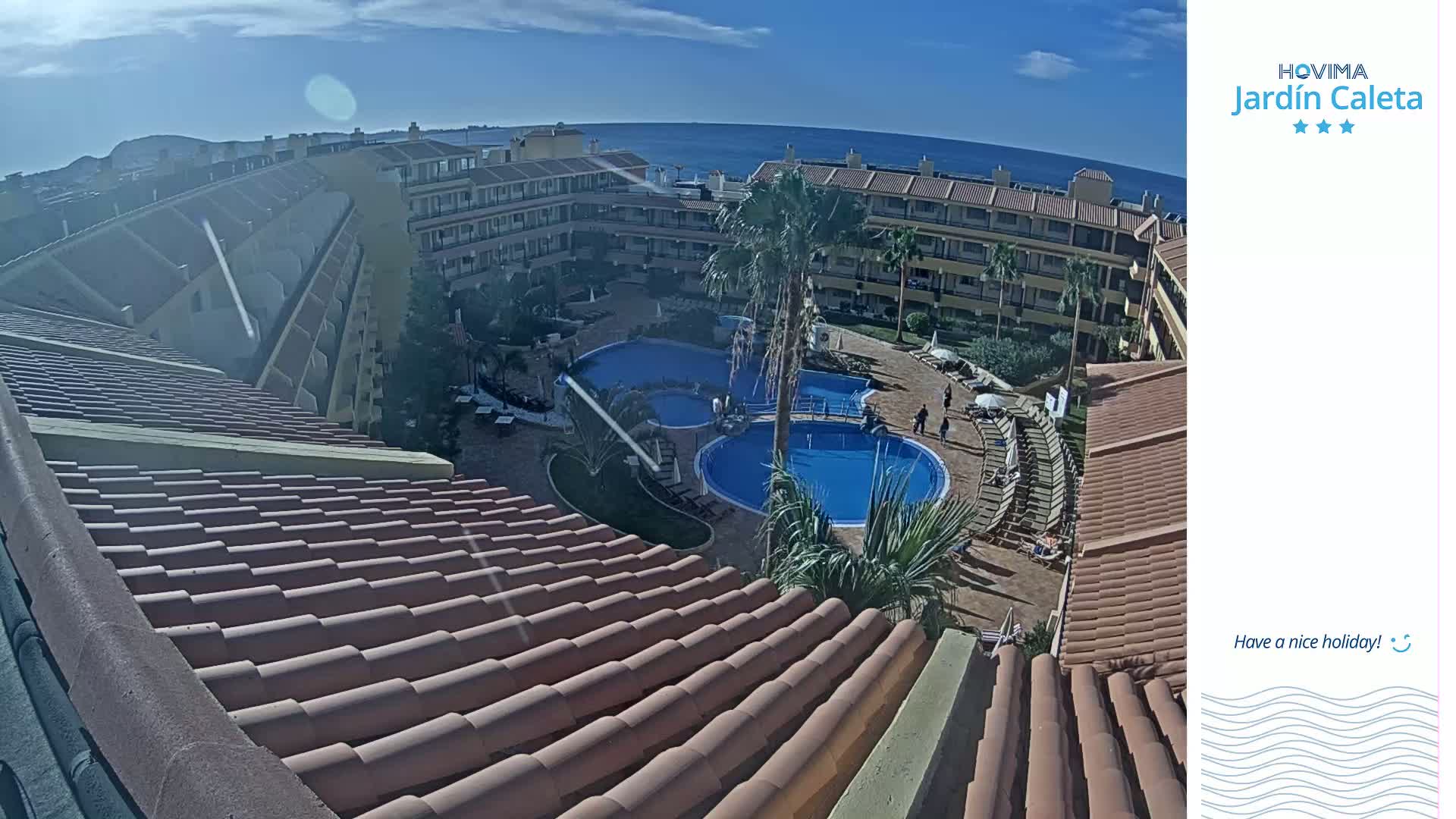 A view from a rooftop overlooking a sunny resort courtyard with two swimming pools and a surrounding complex of buildings near the ocean.