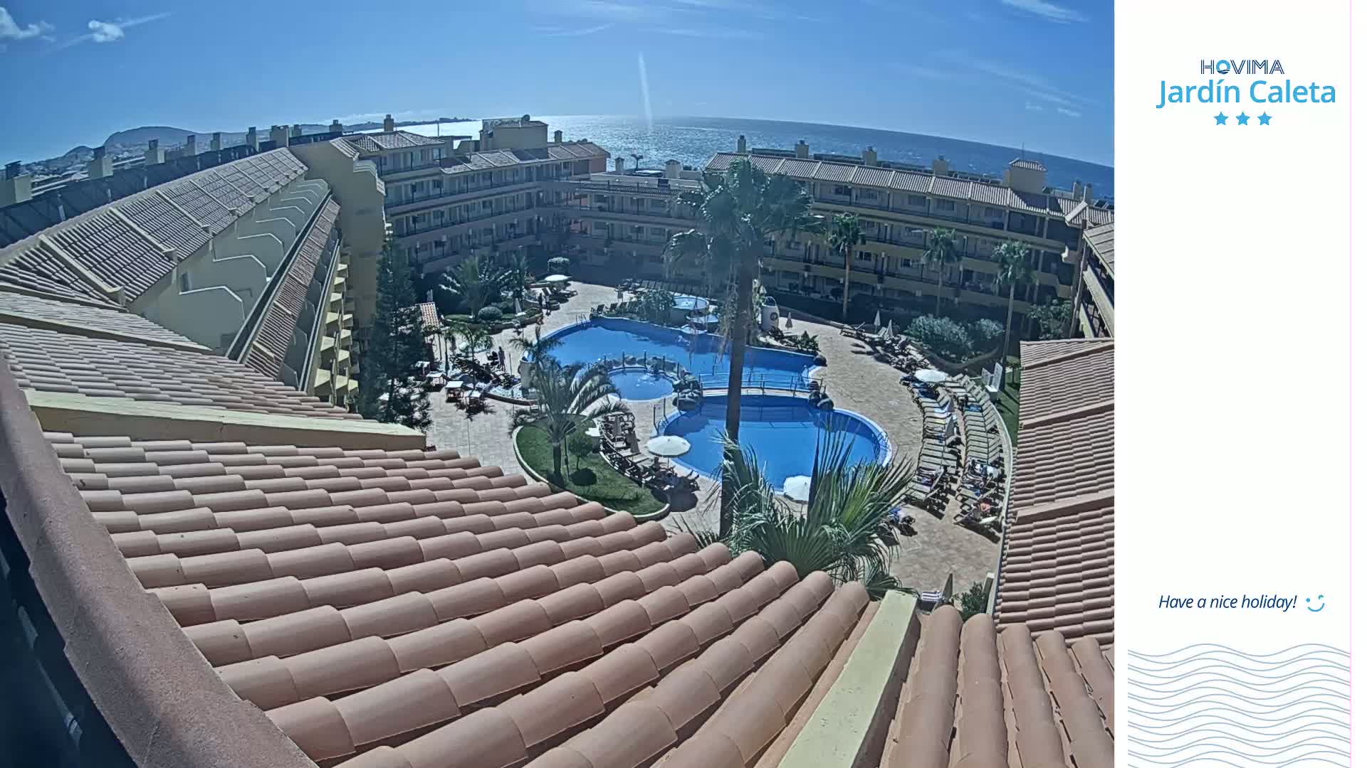 An elevated view of a resort complex with multiple pools surrounded by buildings, under a sunny sky and overlooking a calm ocean.