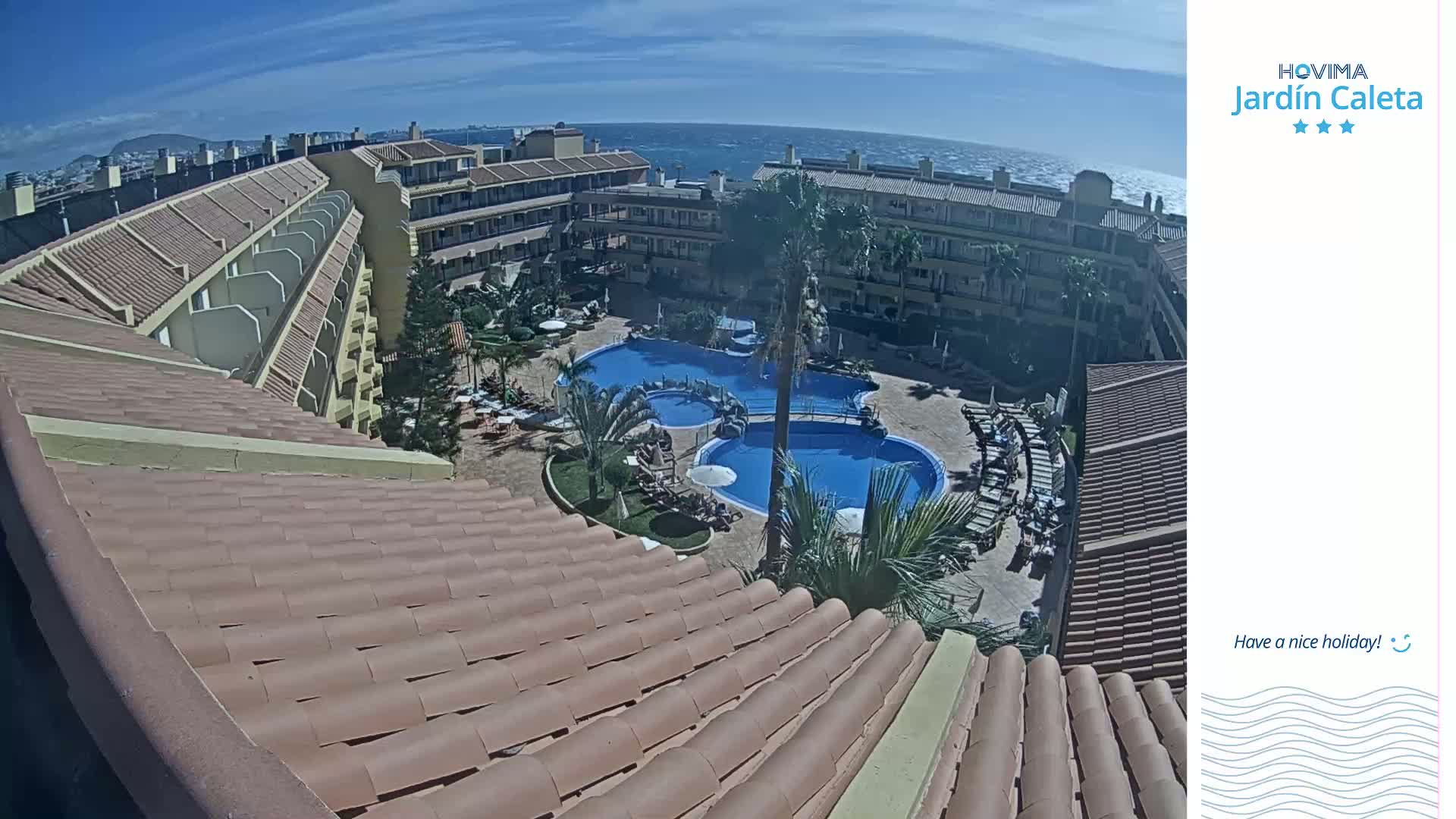 A resort with multiple buildings surrounding a pool complex overlooks the ocean on a sunny day.