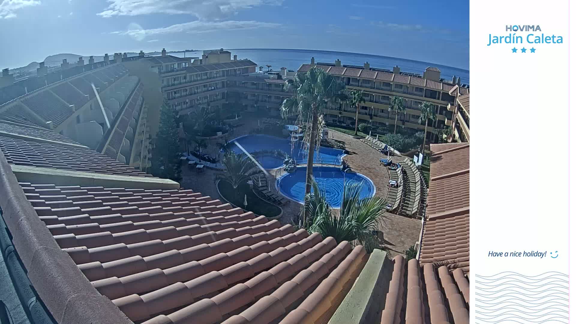 An aerial view of a resort complex with multiple swimming pools, surrounding buildings, and palm trees, overlooking a calm ocean under a mostly sunny sky.