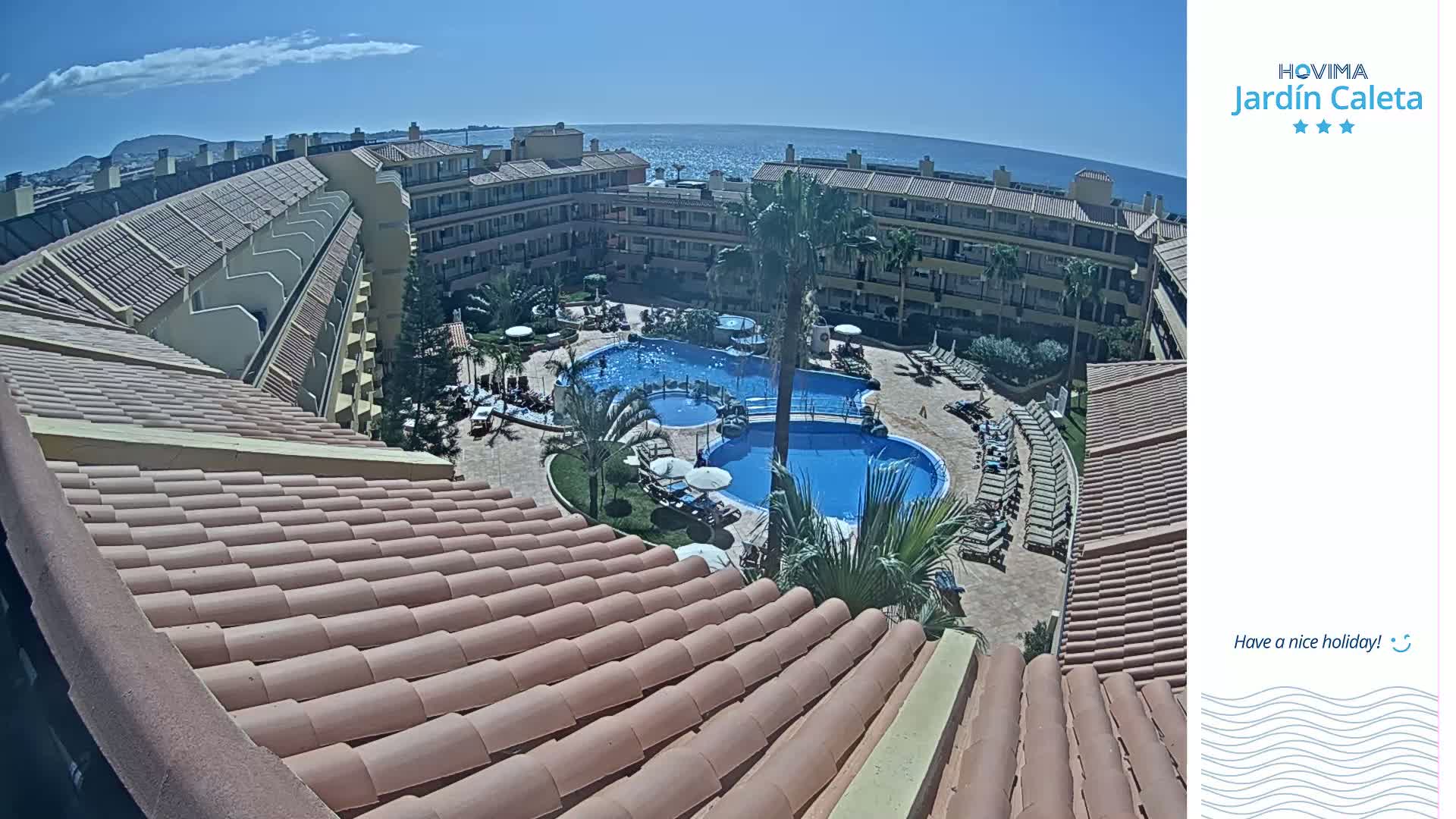 An aerial view of a resort complex with multiple buildings surrounding a large, multi-section swimming pool on a sunny day.