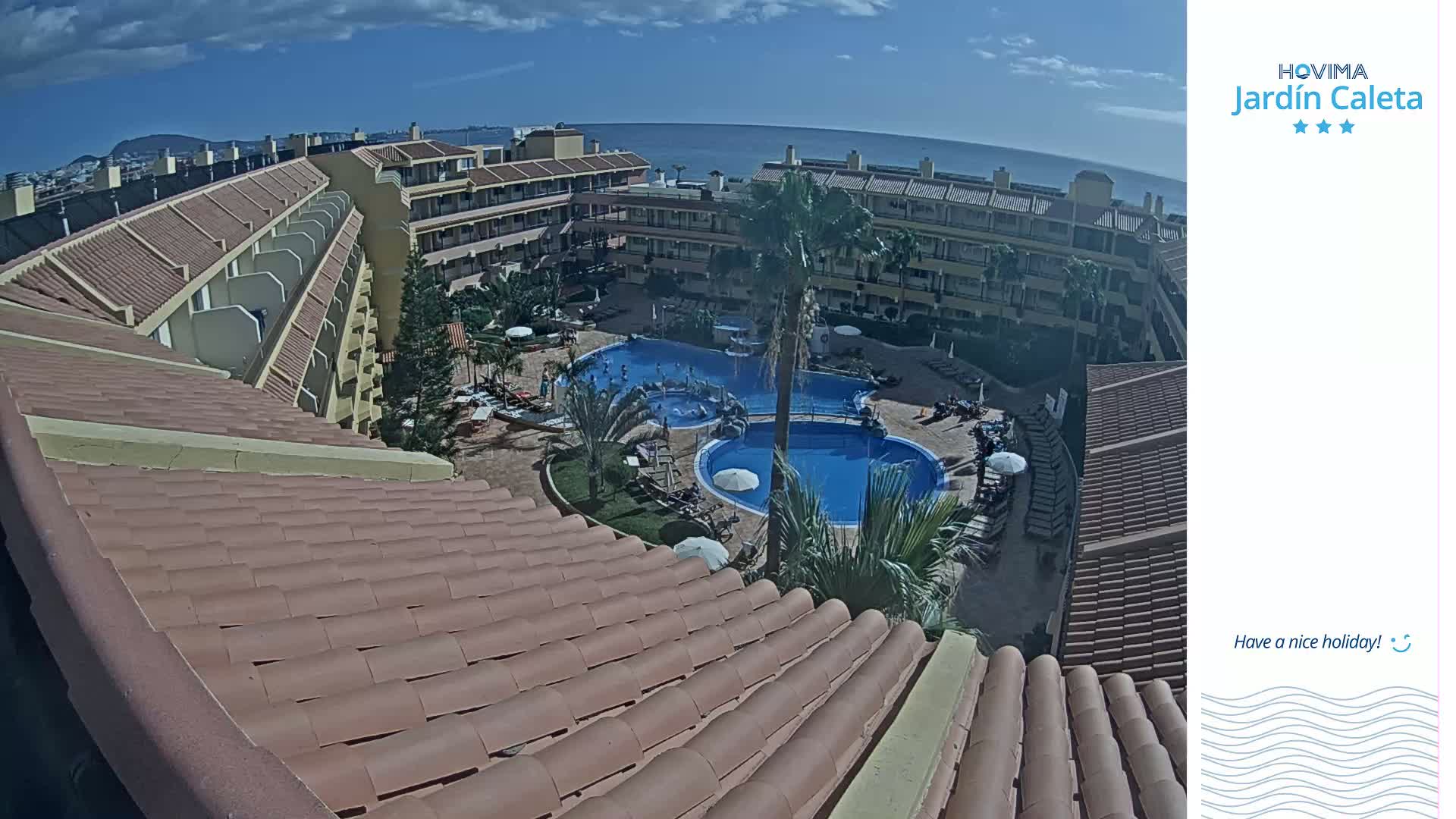 An overhead view shows a resort with a swimming pool and surrounding buildings under a partly cloudy sky.