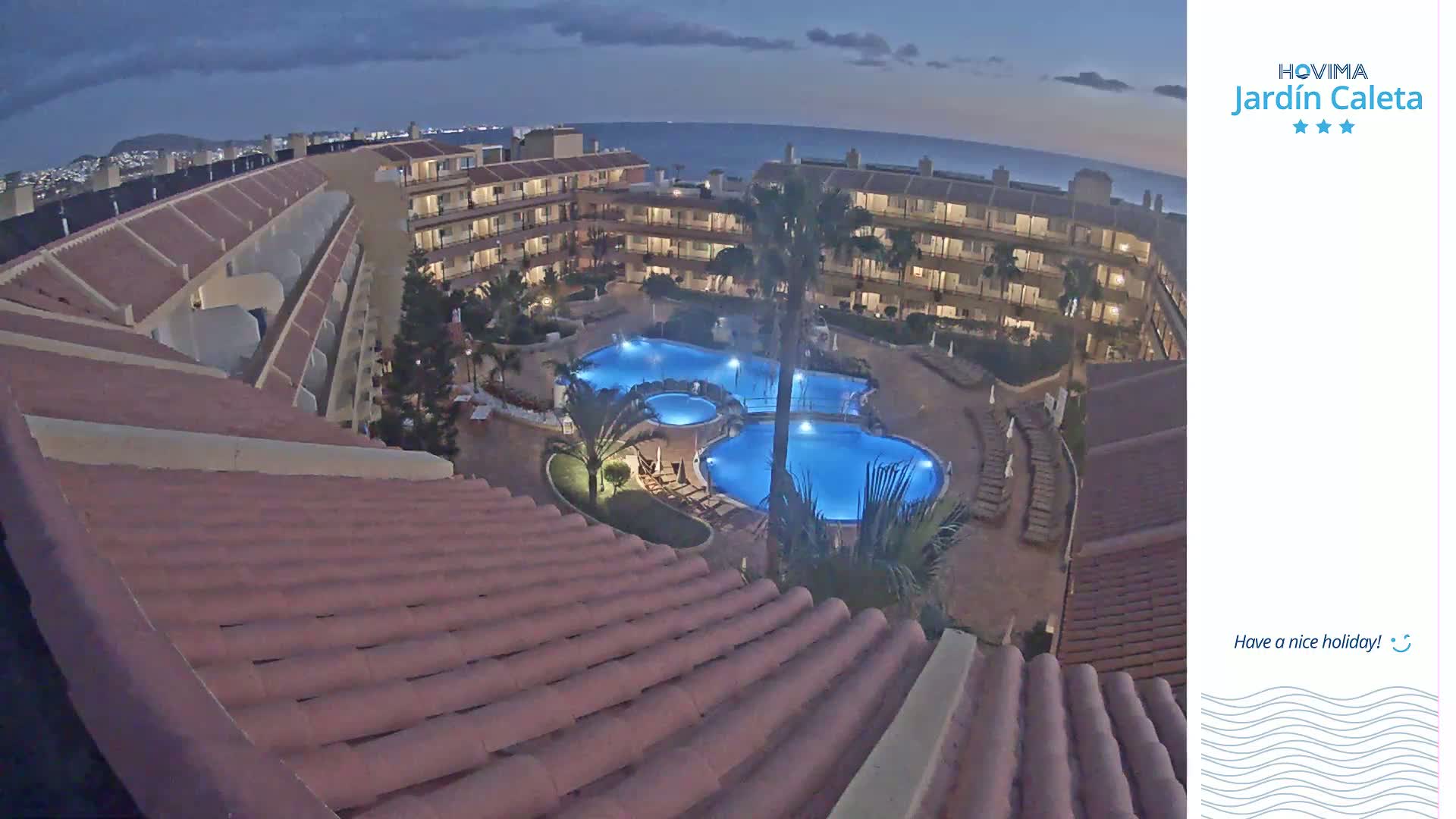 An illuminated, multi-level apartment complex with a large, blue-lit pool at night, overlooking a dark ocean under a mostly clear twilight sky.