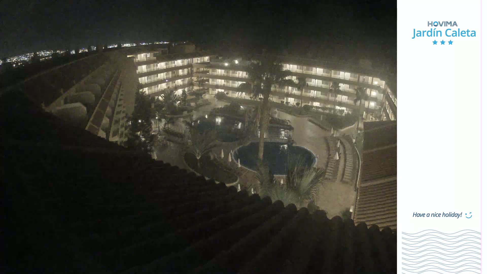 A nighttime aerial view shows a multi-story resort building surrounding a courtyard with pools and landscaping.