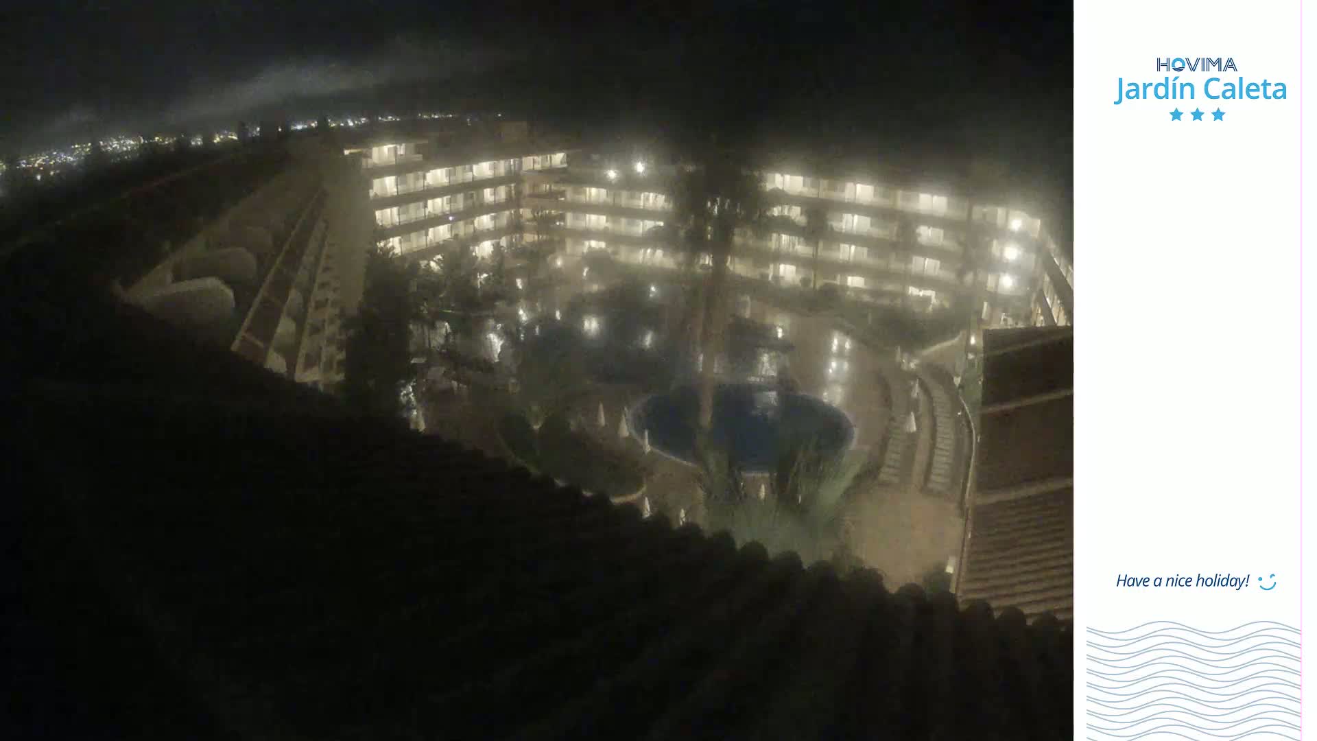 A nighttime, slightly hazy view from above shows a multi-story building complex surrounding a pool area.