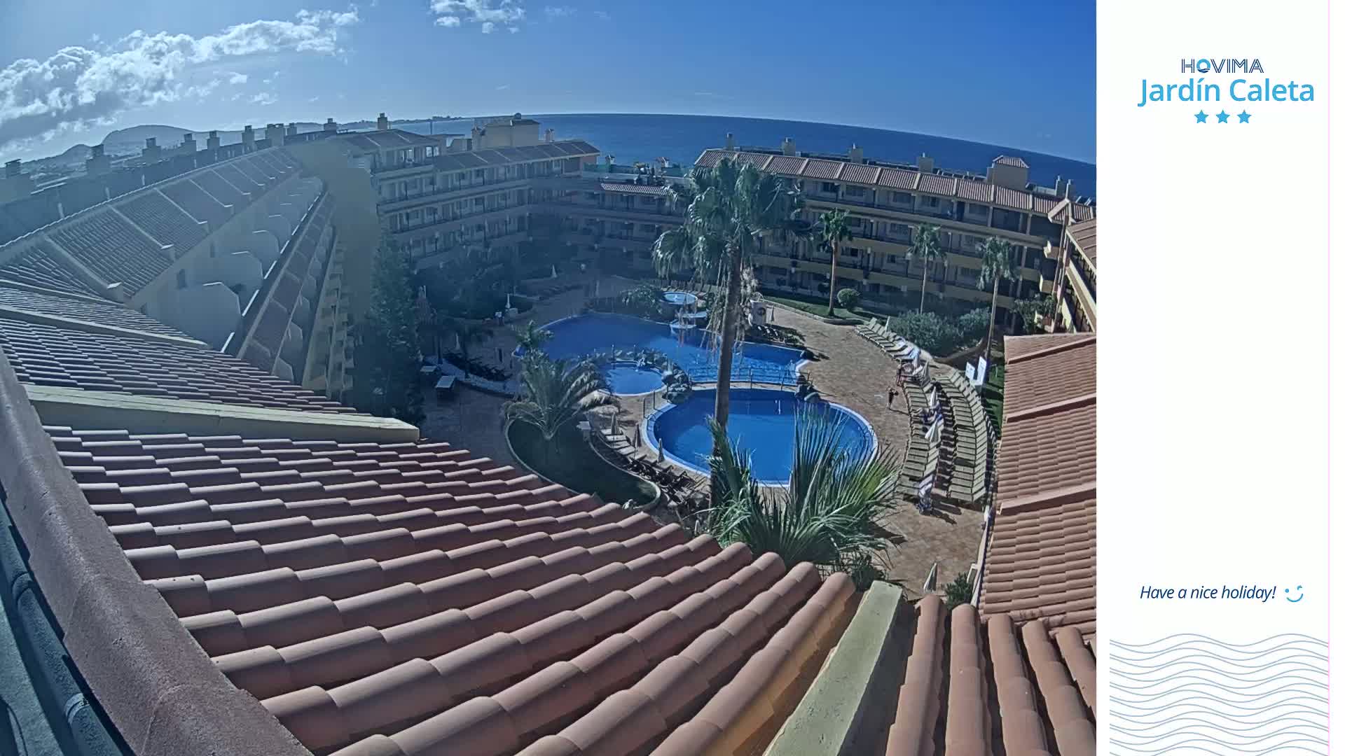 An overhead view shows a resort complex with multiple buildings, a swimming pool, and palm trees, under a mostly sunny sky with some clouds.