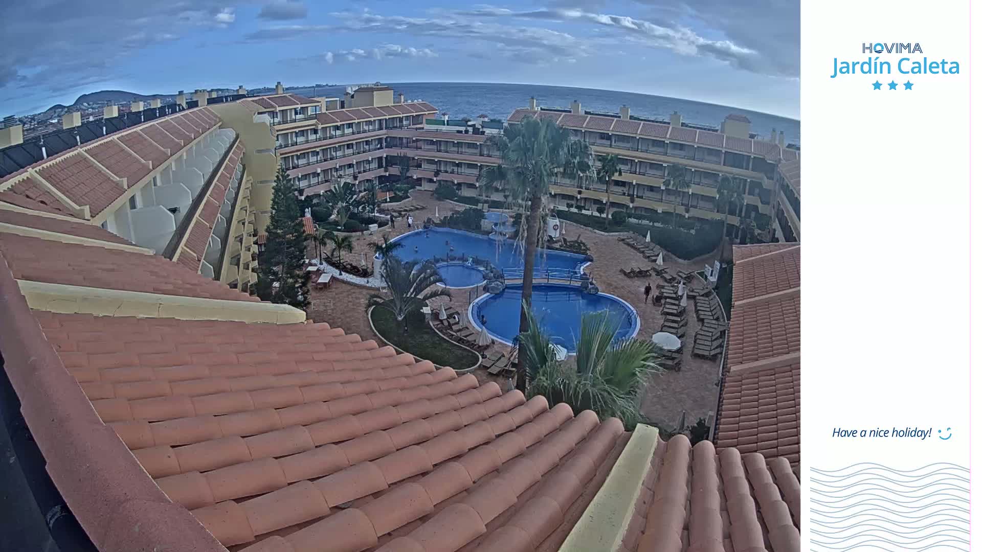 An overhead view shows a resort complex with multiple buildings surrounding a pool area, all under a partly cloudy sky with a calm ocean in the background.