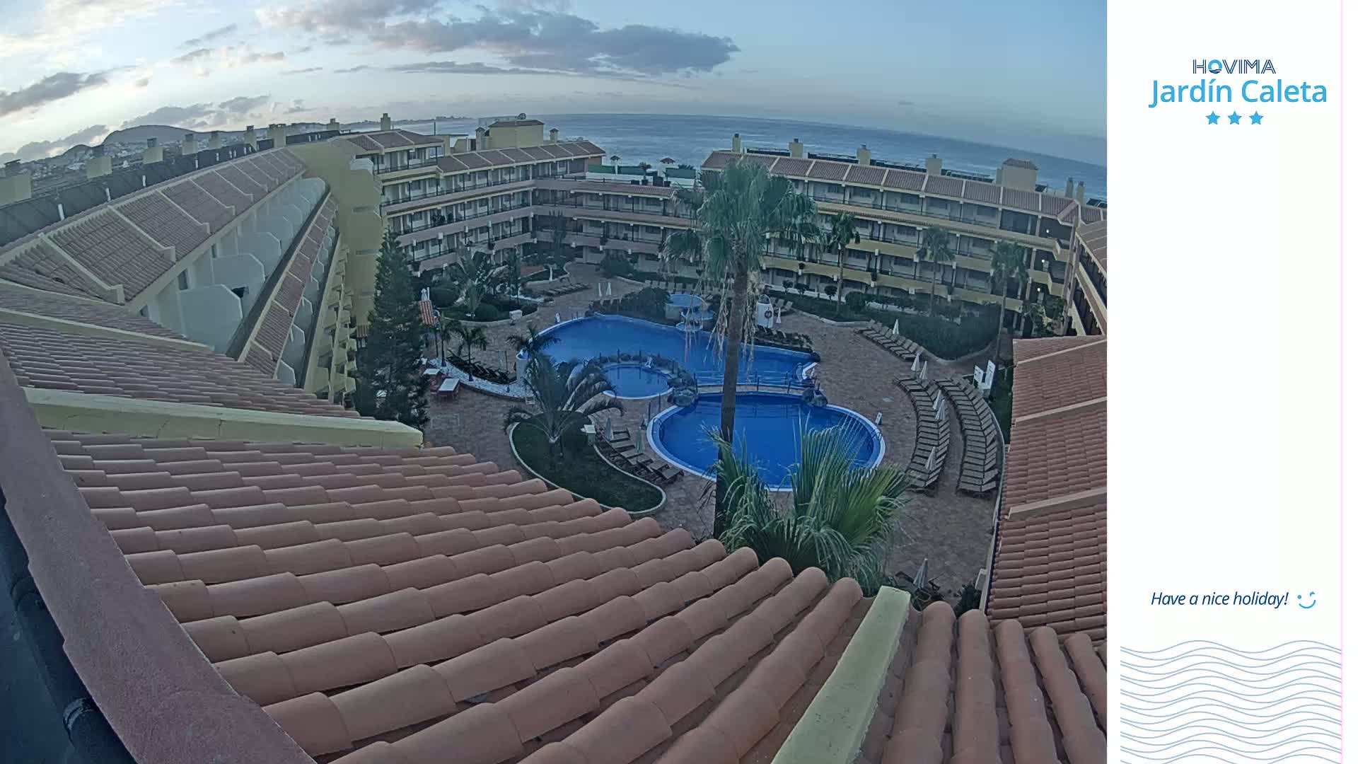 An aerial view shows a resort with multiple buildings surrounding a pool complex, overlooking a calm ocean under a cloudy sky.