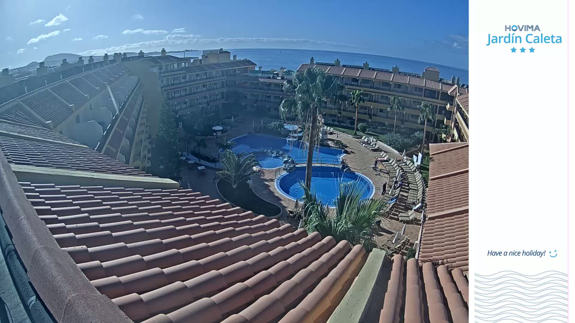 A sunny day overlooking a resort complex with multiple swimming pools, surrounded by buildings and palm trees, and a clear view of the ocean in the distance.