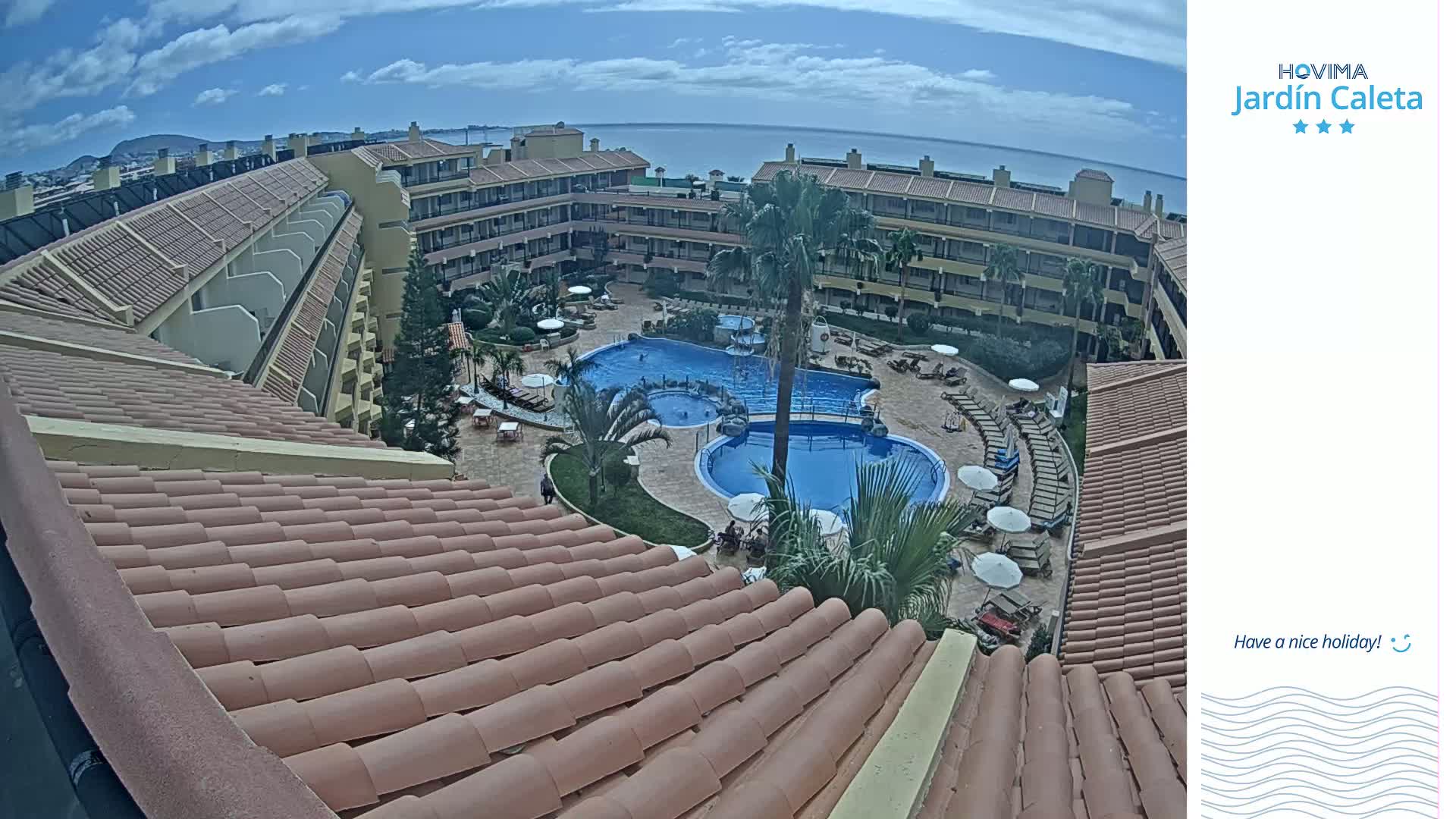 A tiled roof overlooks a resort courtyard with a swimming pool and sun loungers under a partly cloudy sky.