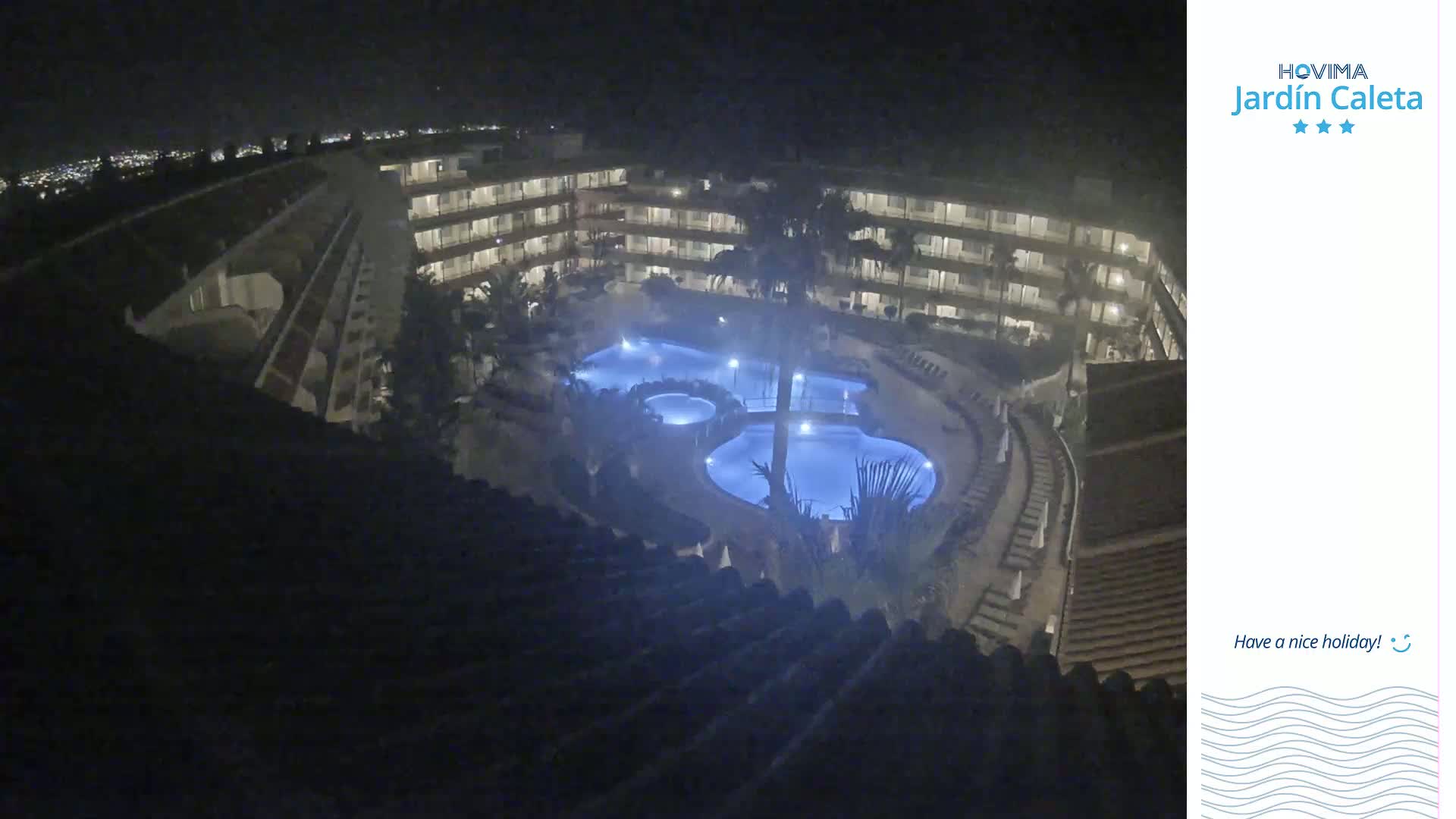 A nighttime, high-angle view shows a multi-story building complex surrounding a lit-up pool area.