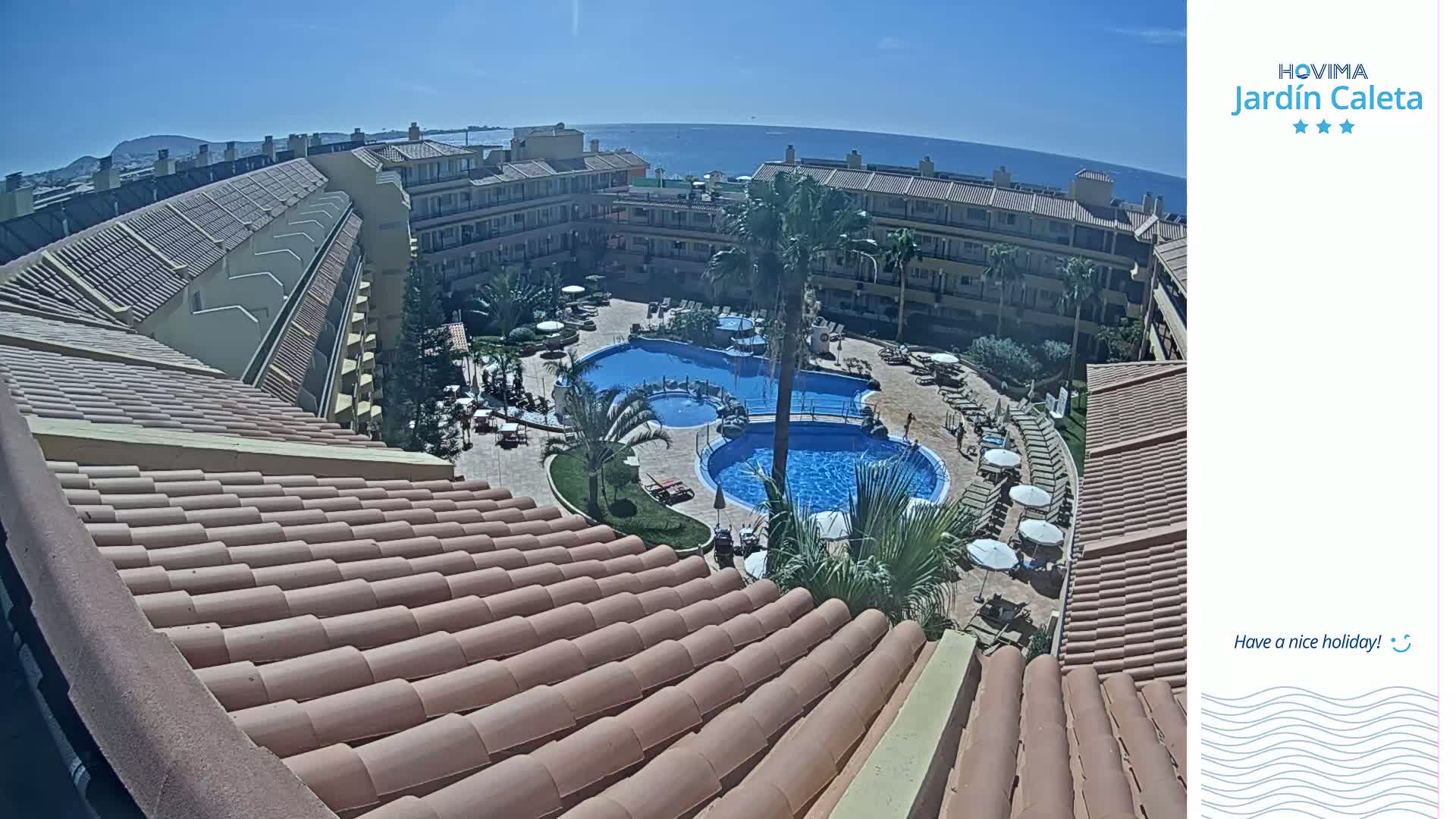 A sunny view from a rooftop shows a resort courtyard with two swimming pools surrounded by buildings and lounge chairs.
