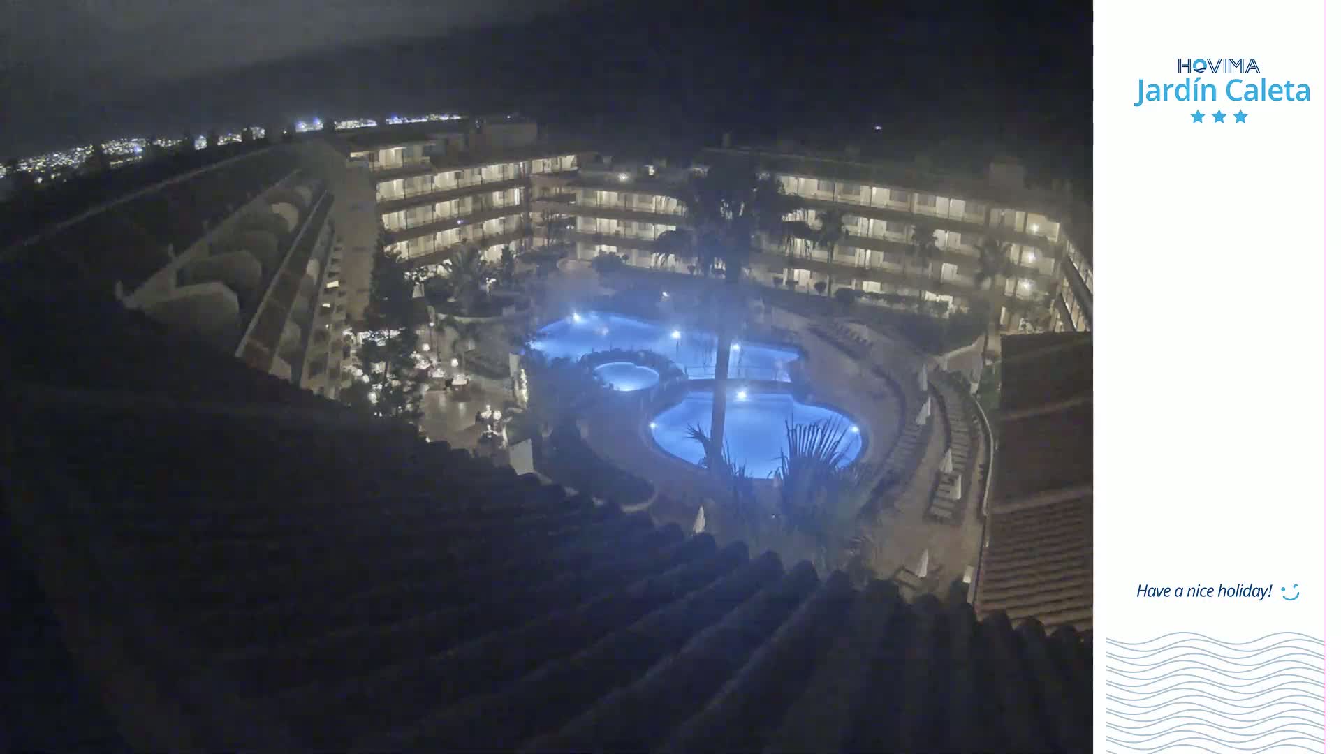 A nighttime view from a rooftop shows a multi-story building complex surrounding a lit swimming pool.