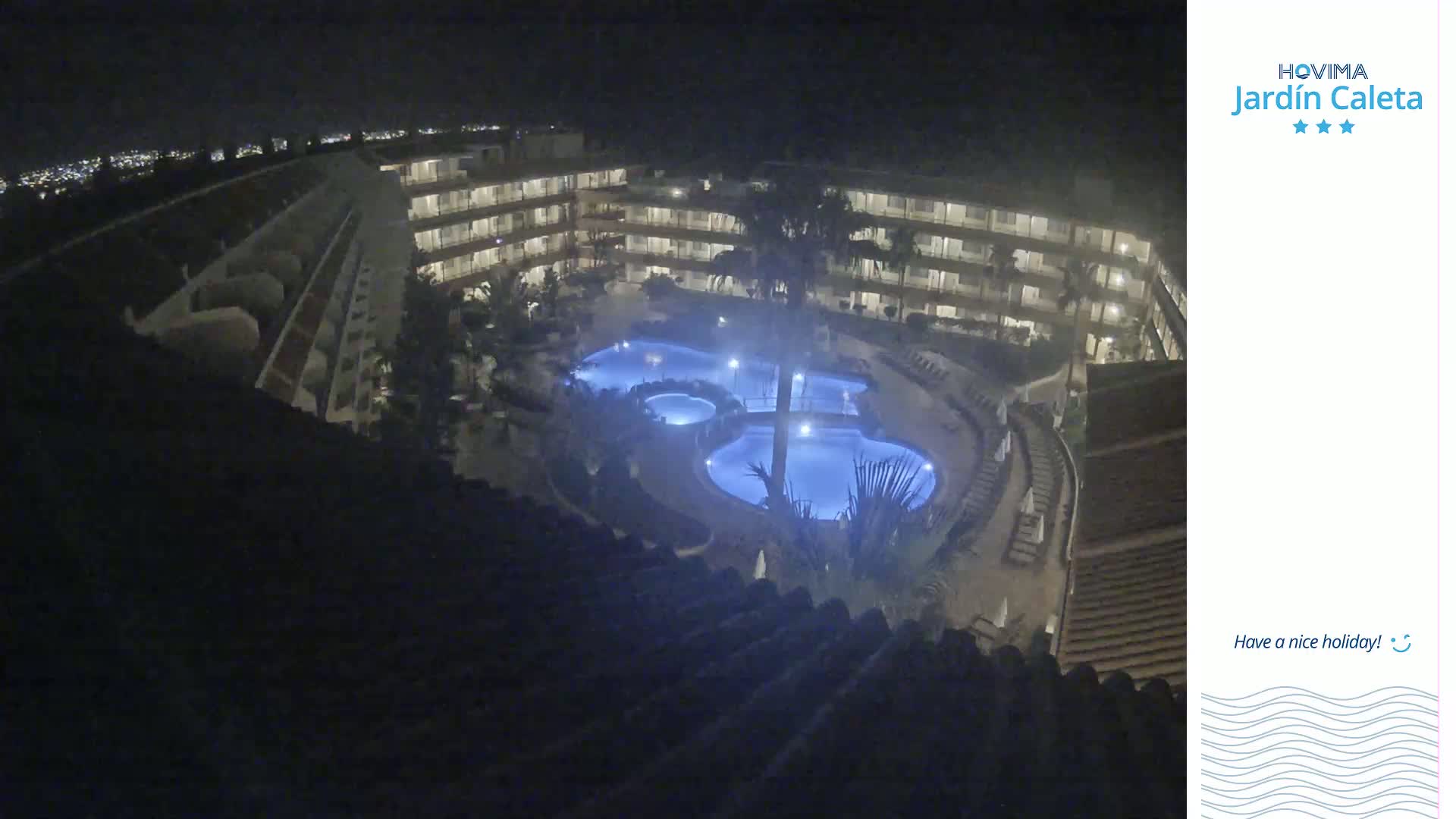 A nighttime aerial view shows a multi-story building surrounding a lit, kidney-shaped swimming pool complex.